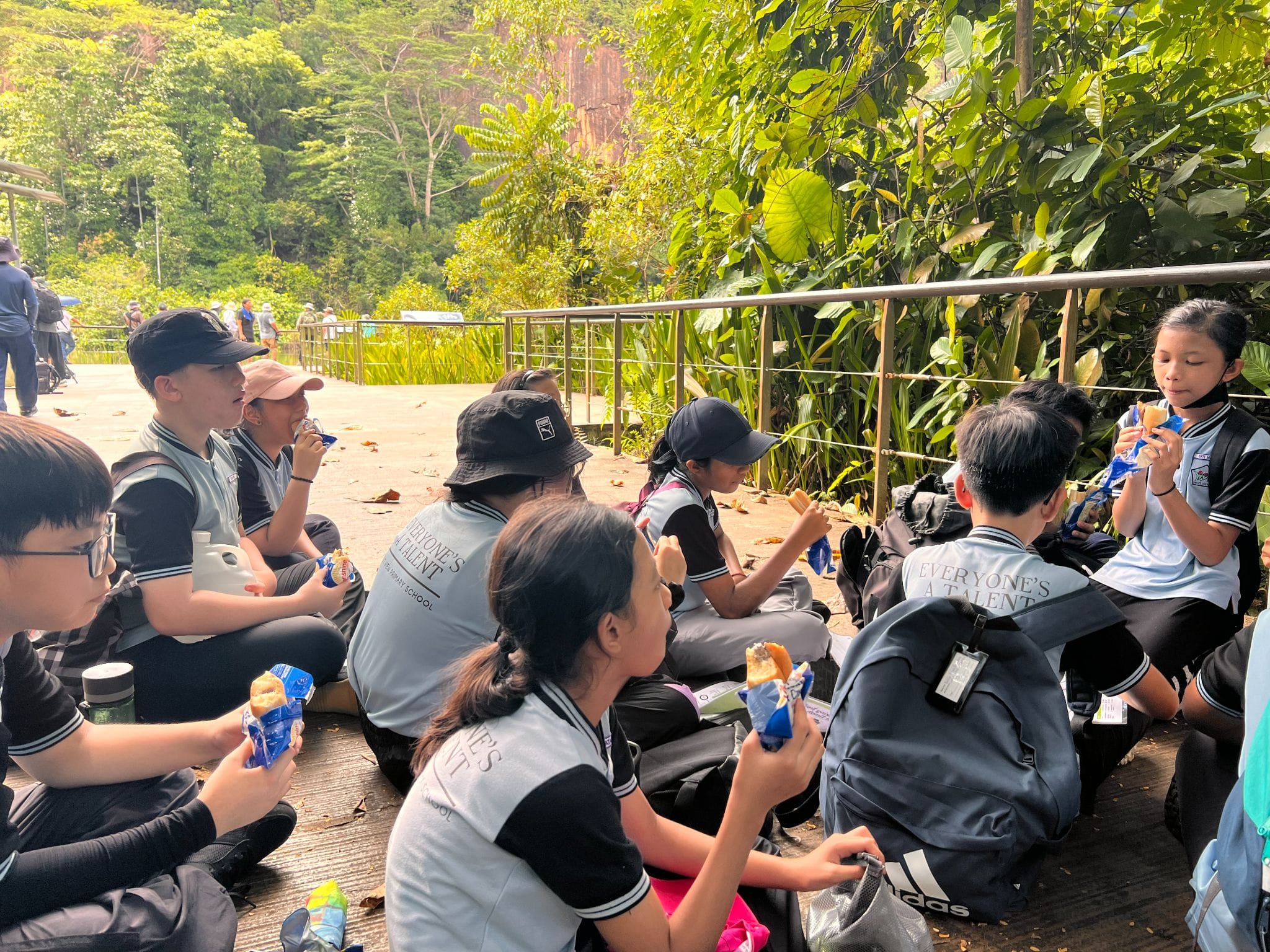 School children in gray uniforms sit on the ground eating snacks outdoors near a railing with green trees behind them.