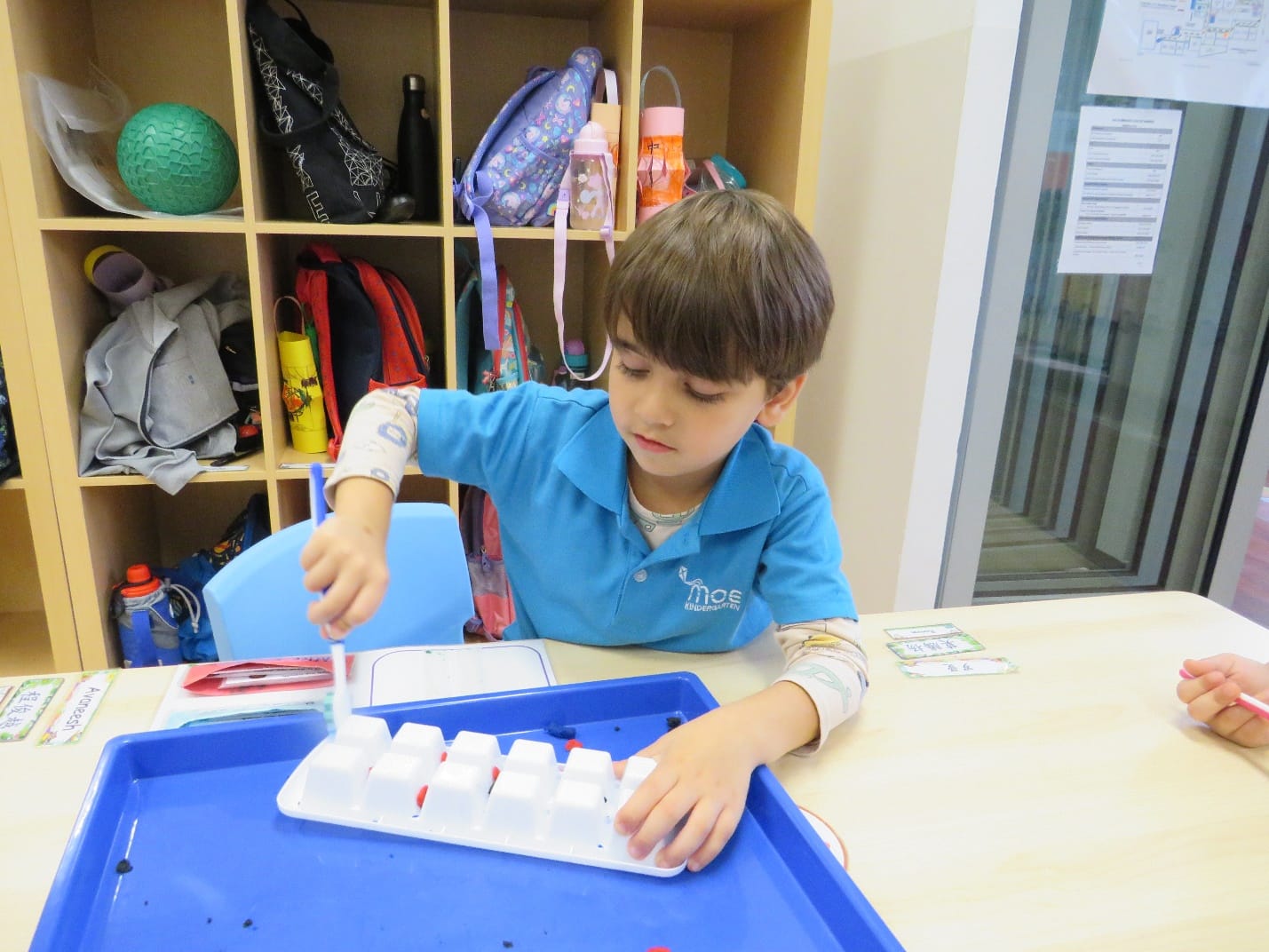 Young boy in a blue school shirt uses a brush on a white tray with small cubes at a classroom desk.