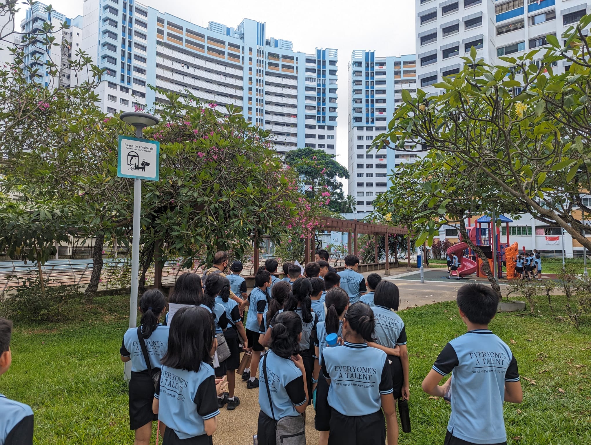Students in blue "Everyone's A Talent" uniforms gather in a park near tall residential buildings and a playground.