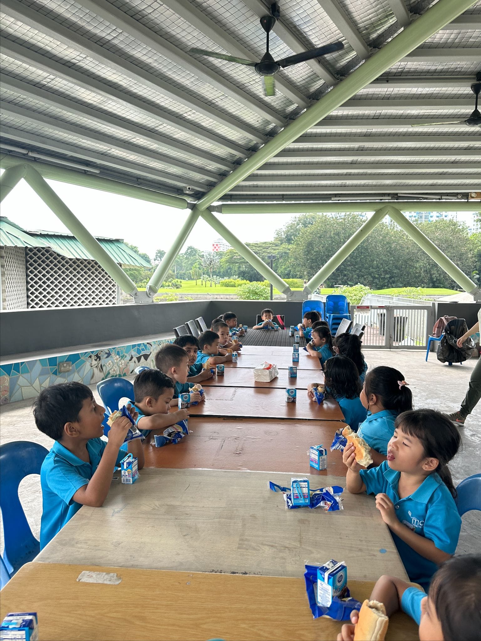 Young children in blue uniforms eat snacks and drink juice boxes at a long wooden table under an open shelter.