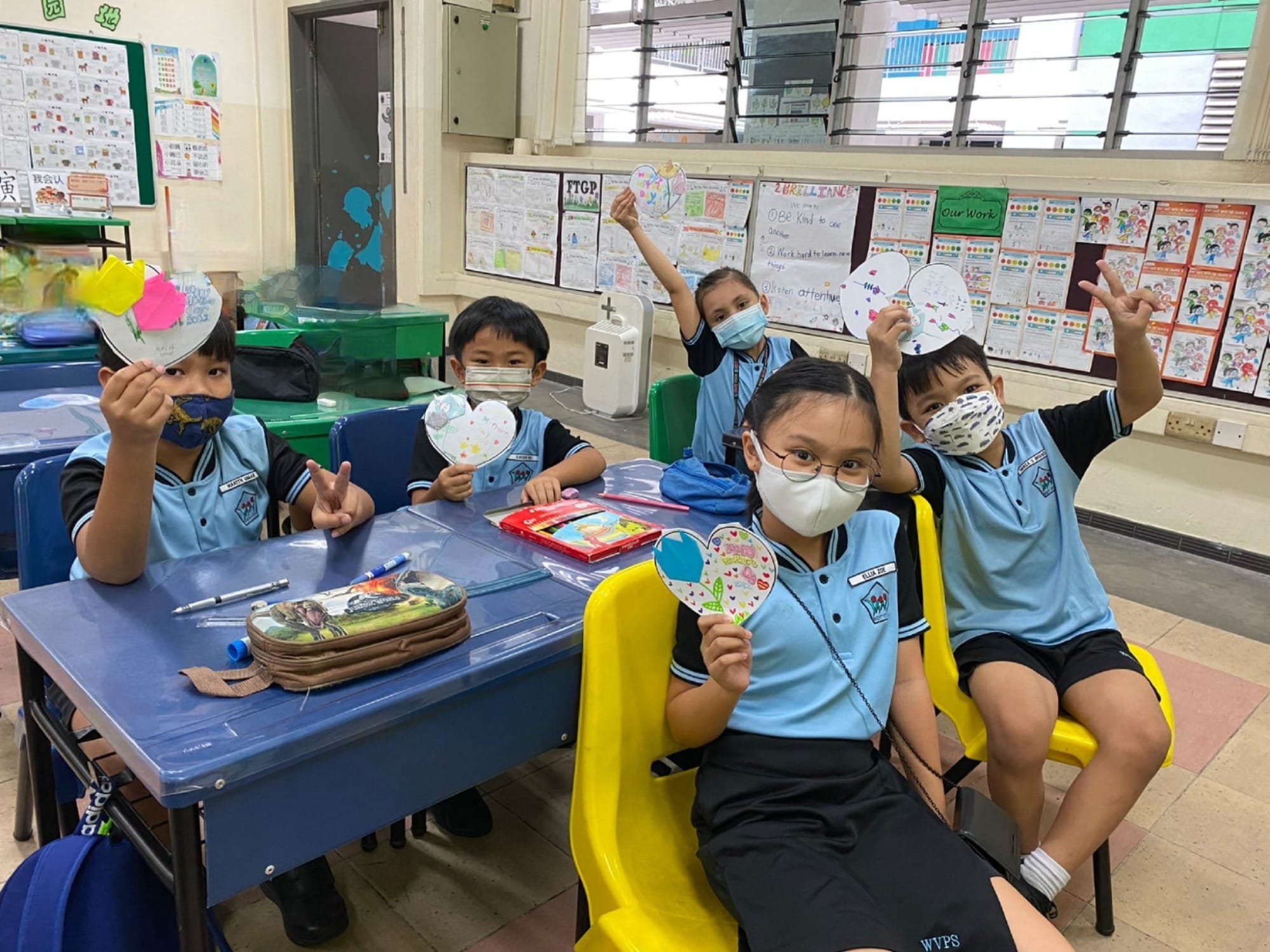 Five young students in blue uniforms and face masks hold up decorated heart-shaped paper crafts in a classroom.