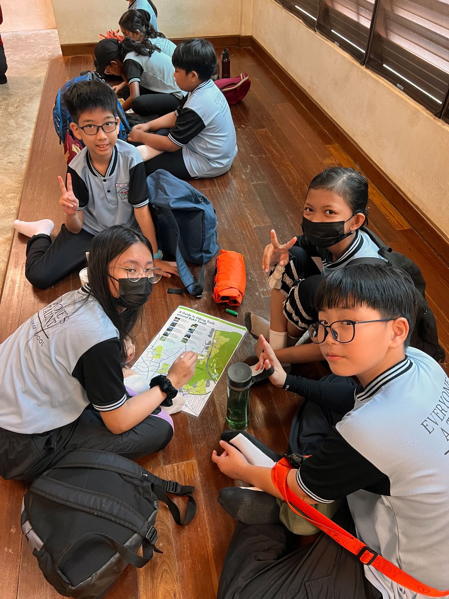 Several school children in uniforms sit on a wooden floor, some wearing masks, looking at a hiking trail map.