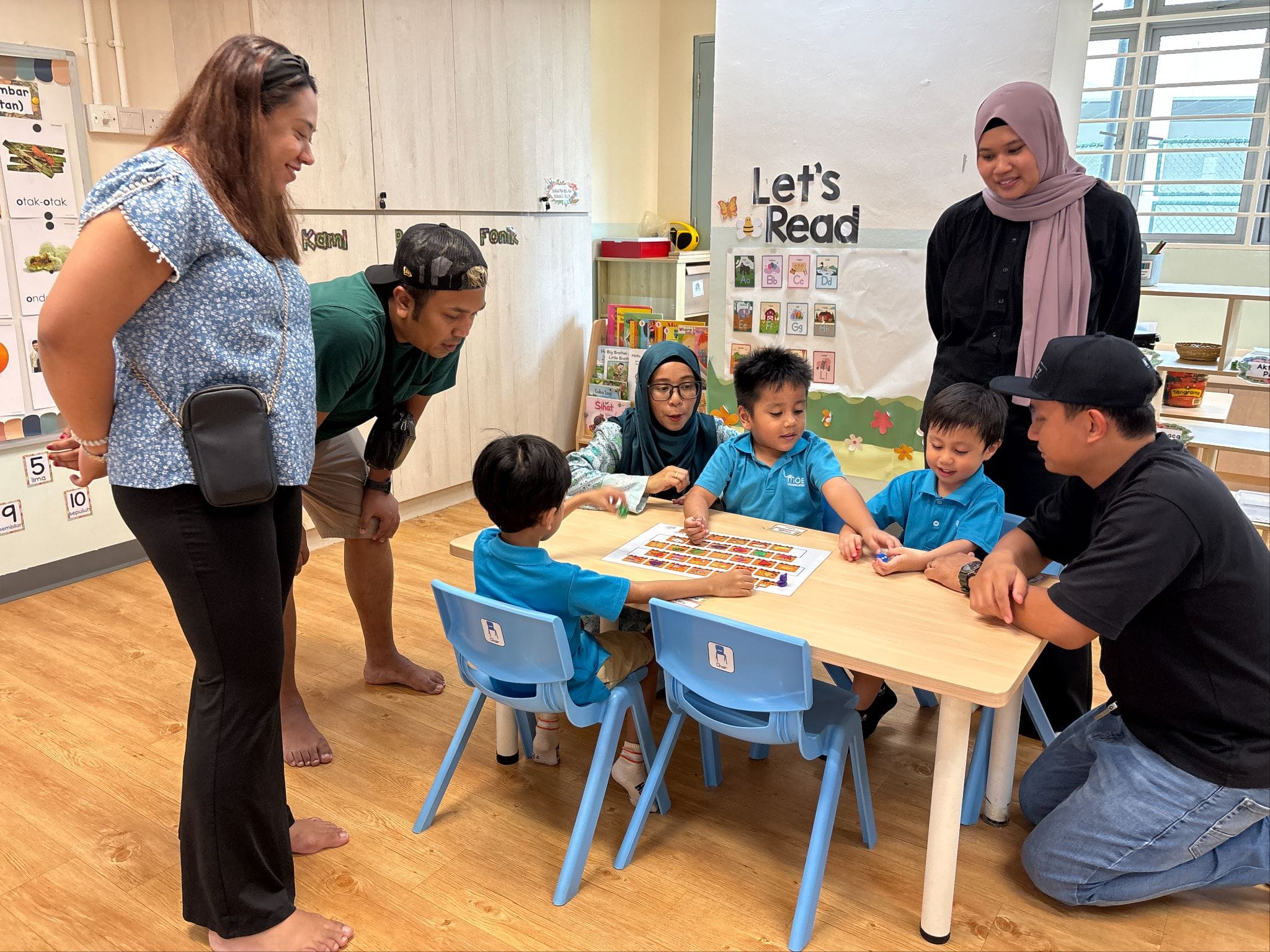 Three young children in blue uniforms sit at a table with adults around them in a bright classroom.
