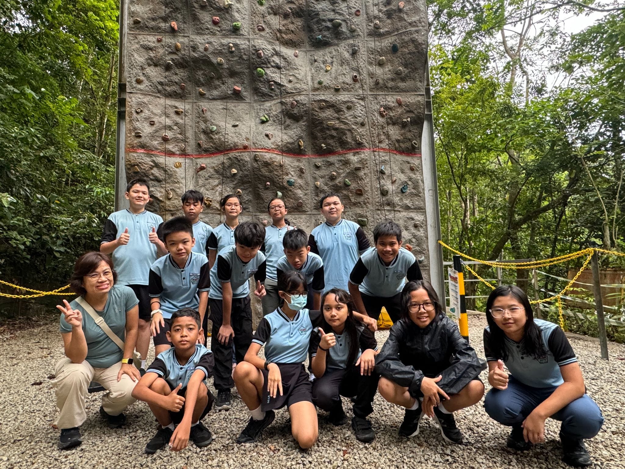 A group of students in light blue uniforms pose in front of an outdoor rock climbing wall surrounded by trees.