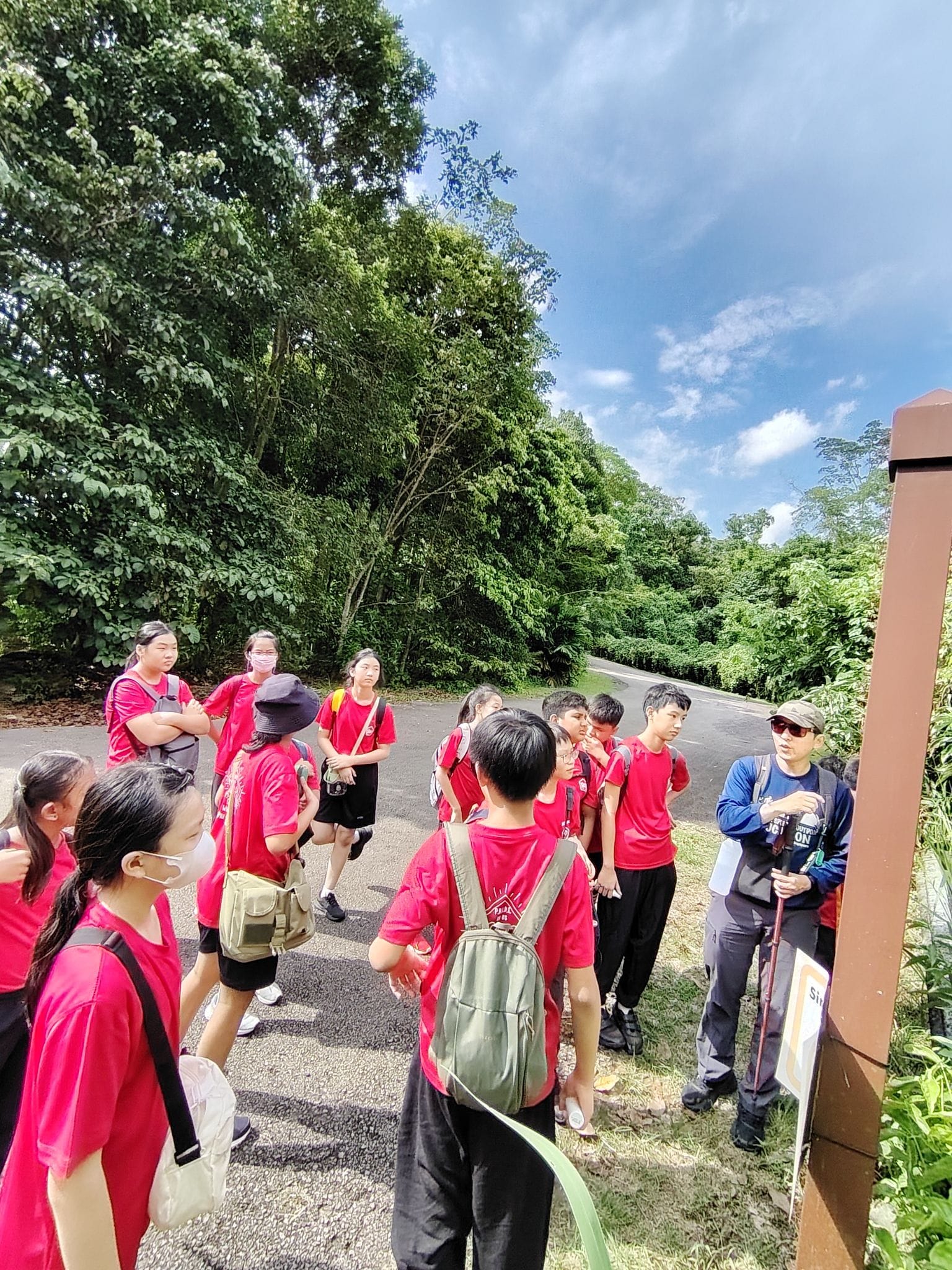 A group of students in red shirts with backpacks listens to a guide outdoors near a forested trail.
