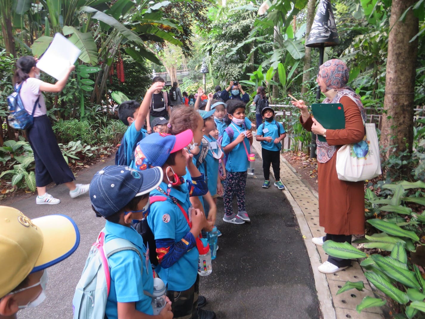 A teacher addresses a group of young children in blue uniforms on a lush green garden path during a field trip.
