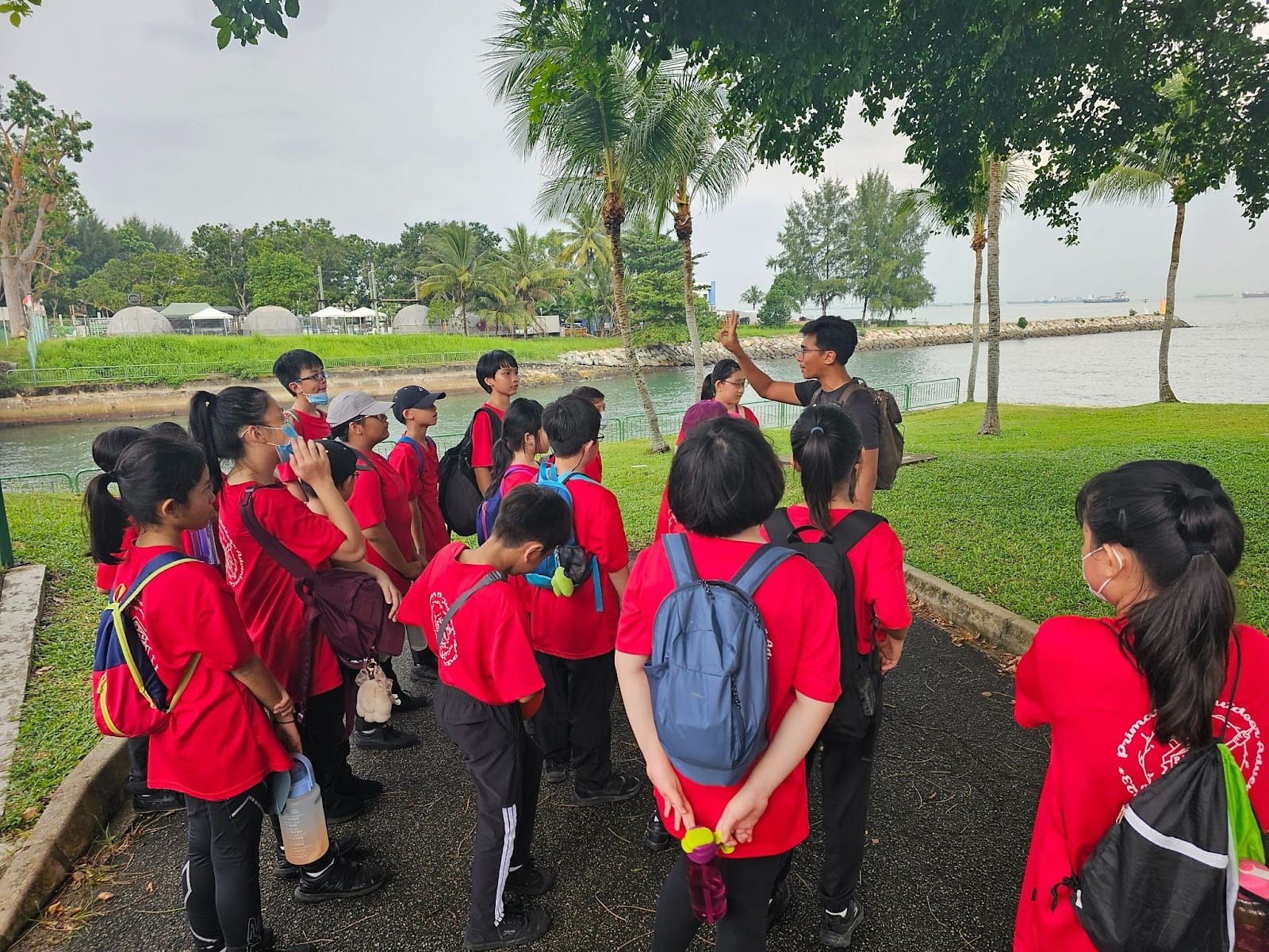 A group of students in red shirts with backpacks listens to a guide near a waterfront park with palm trees.