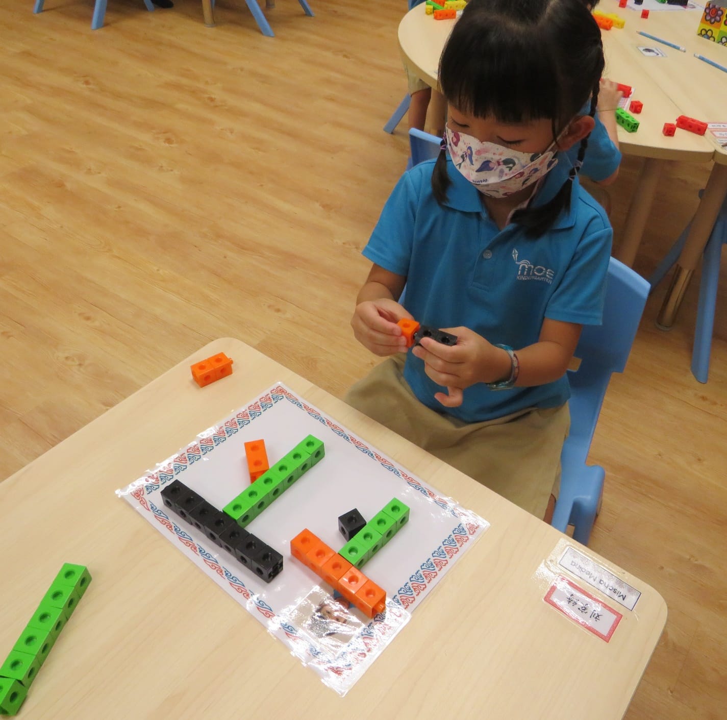A young masked child in a blue shirt arranges colorful linking cubes on a paper at a classroom desk.