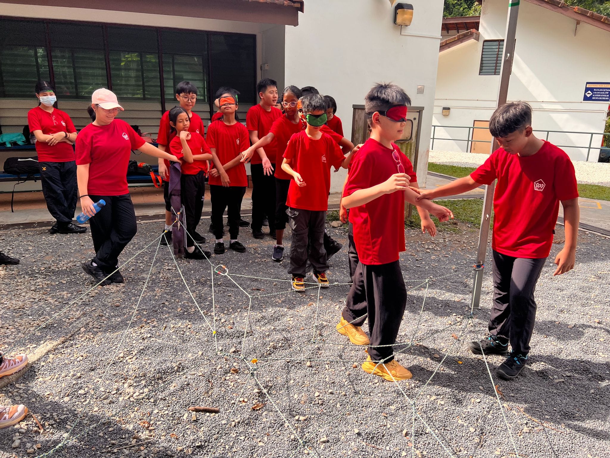 Children in red shirts do a blindfolded team-building activity navigating a rope obstacle course on gravel outdoors.