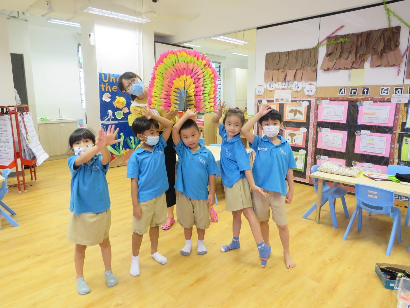 Five young children in blue uniforms pose with raised hands in a colorful classroom with an art gallery display.