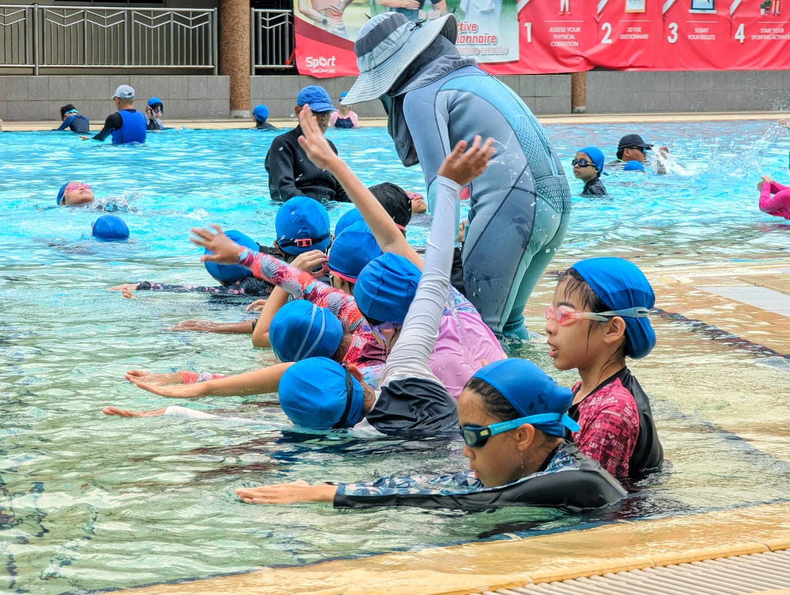 Children in blue swim caps practice arm movements in a pool while an instructor in a sun hat guides them.