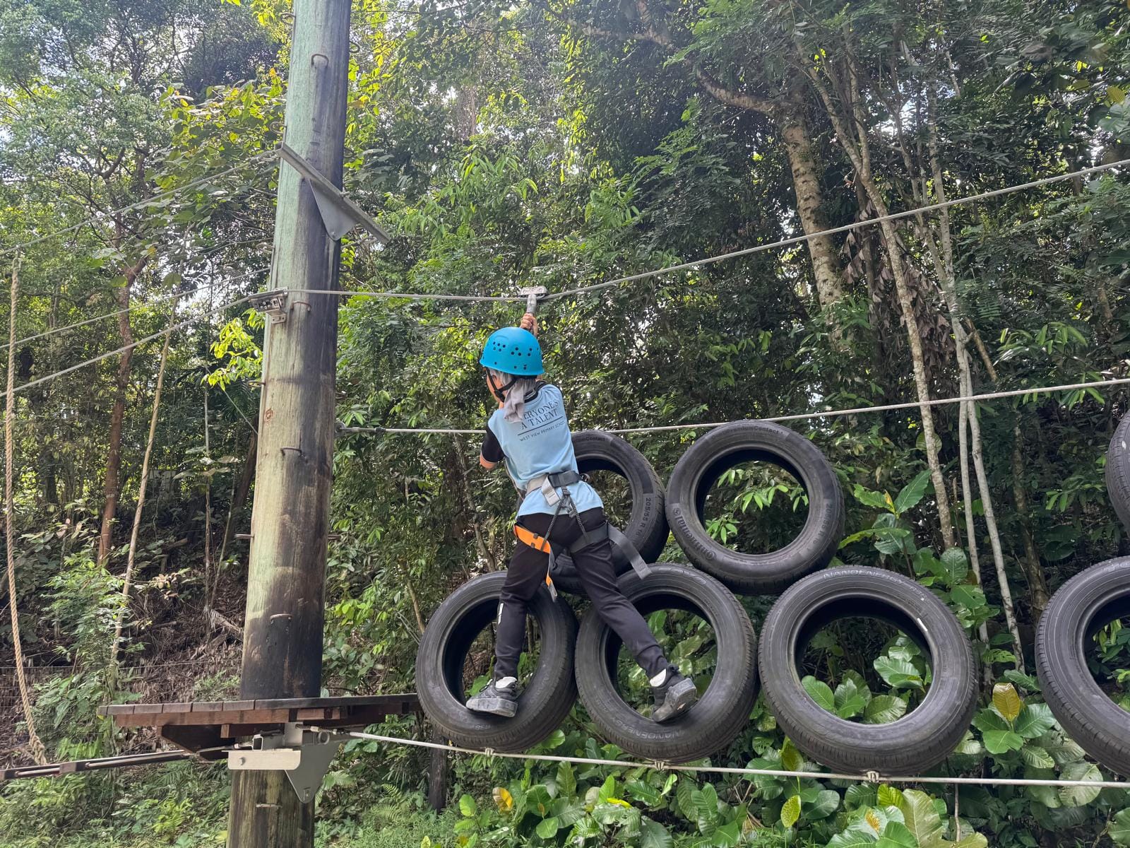 Child in blue helmet and harness navigating a rope course obstacle made of suspended tires in a forest.