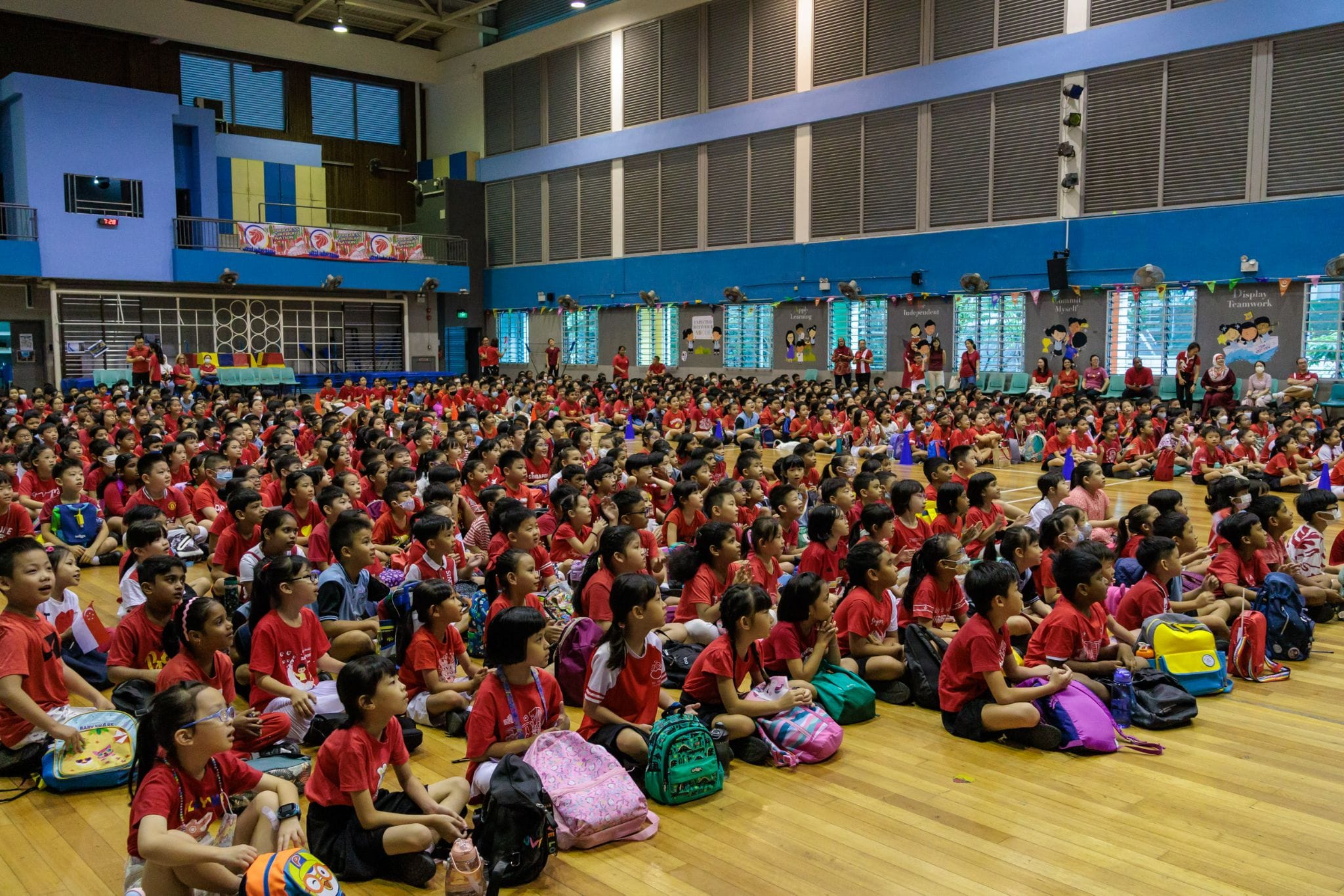 Hundreds of children in red shirts sit on a school gymnasium floor with backpacks during an assembly.