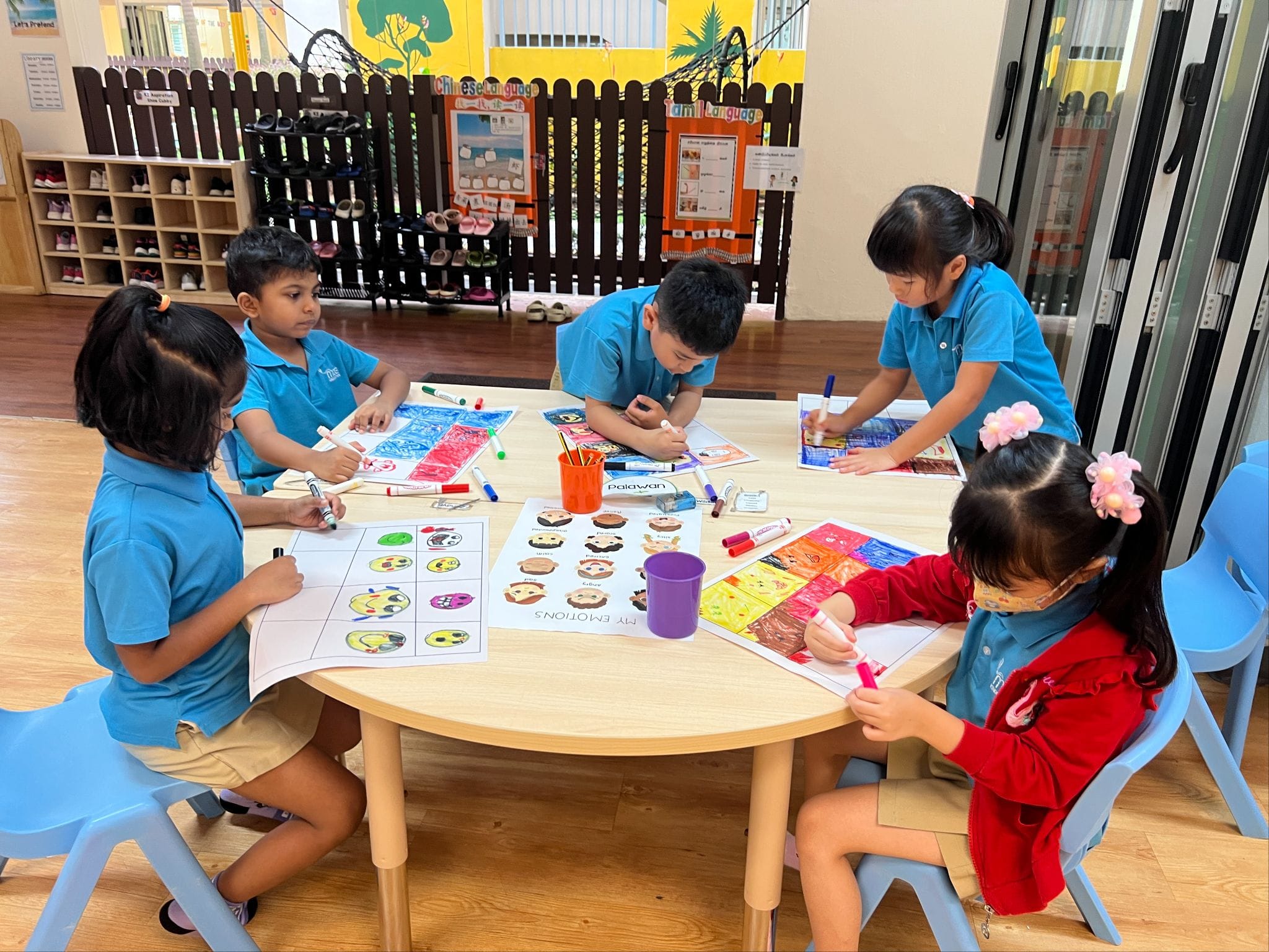 Five young children in blue uniforms sit around a round table coloring worksheets with markers in a classroom.