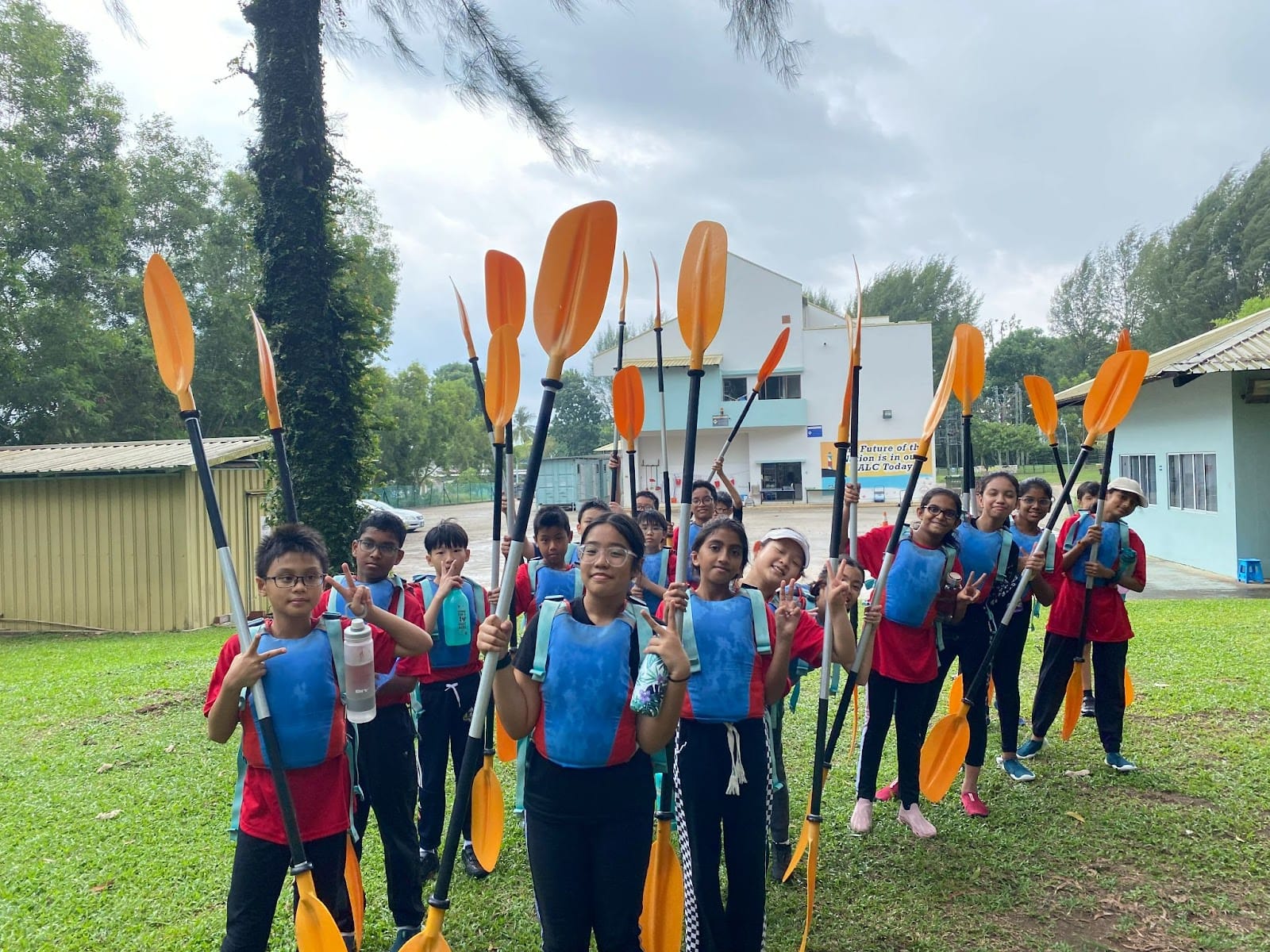 A group of children in blue life vests holding orange kayak paddles up, posing outdoors near a building.