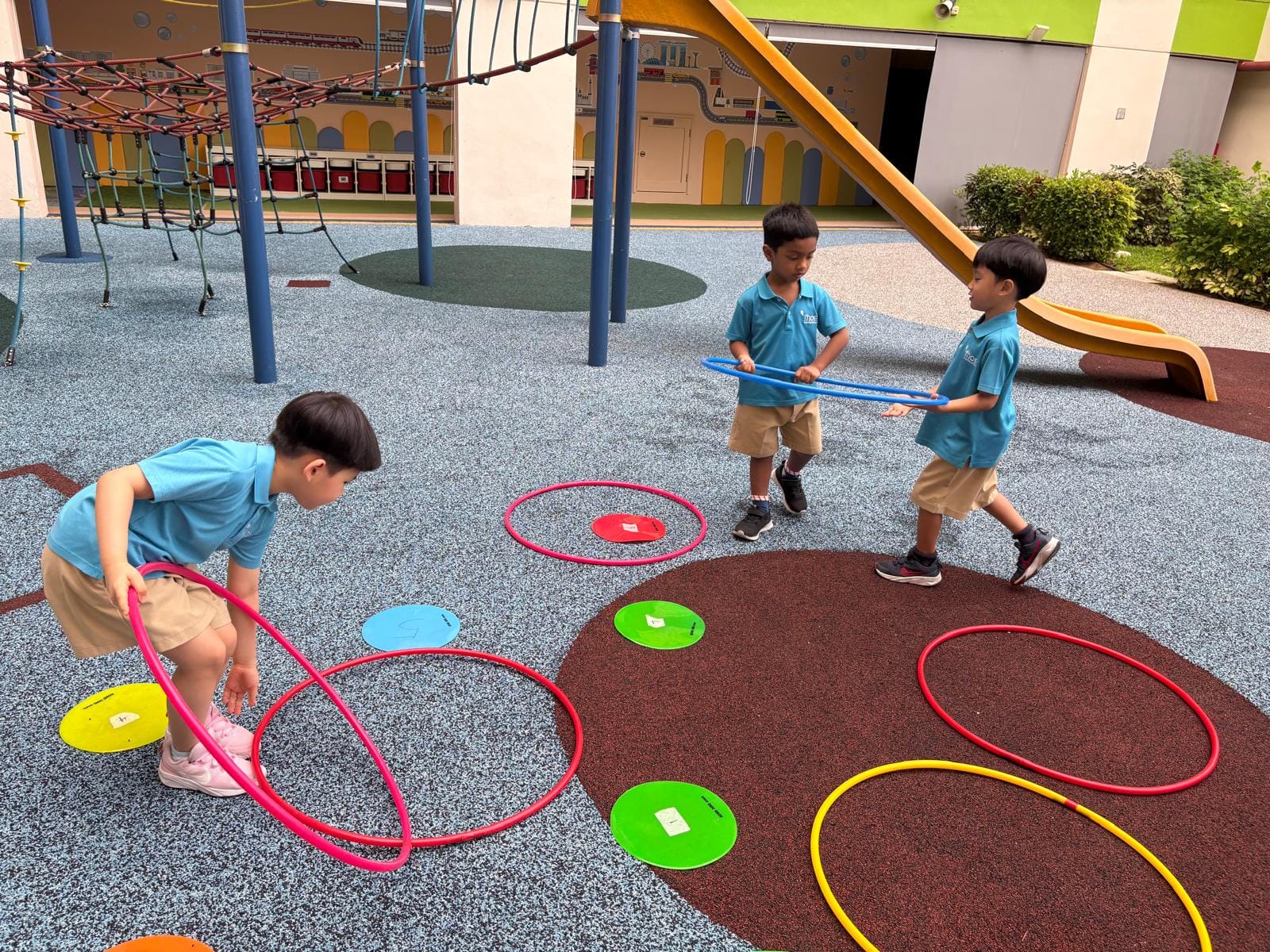 Three young children in blue uniforms play with colorful hula hoops on a school playground.