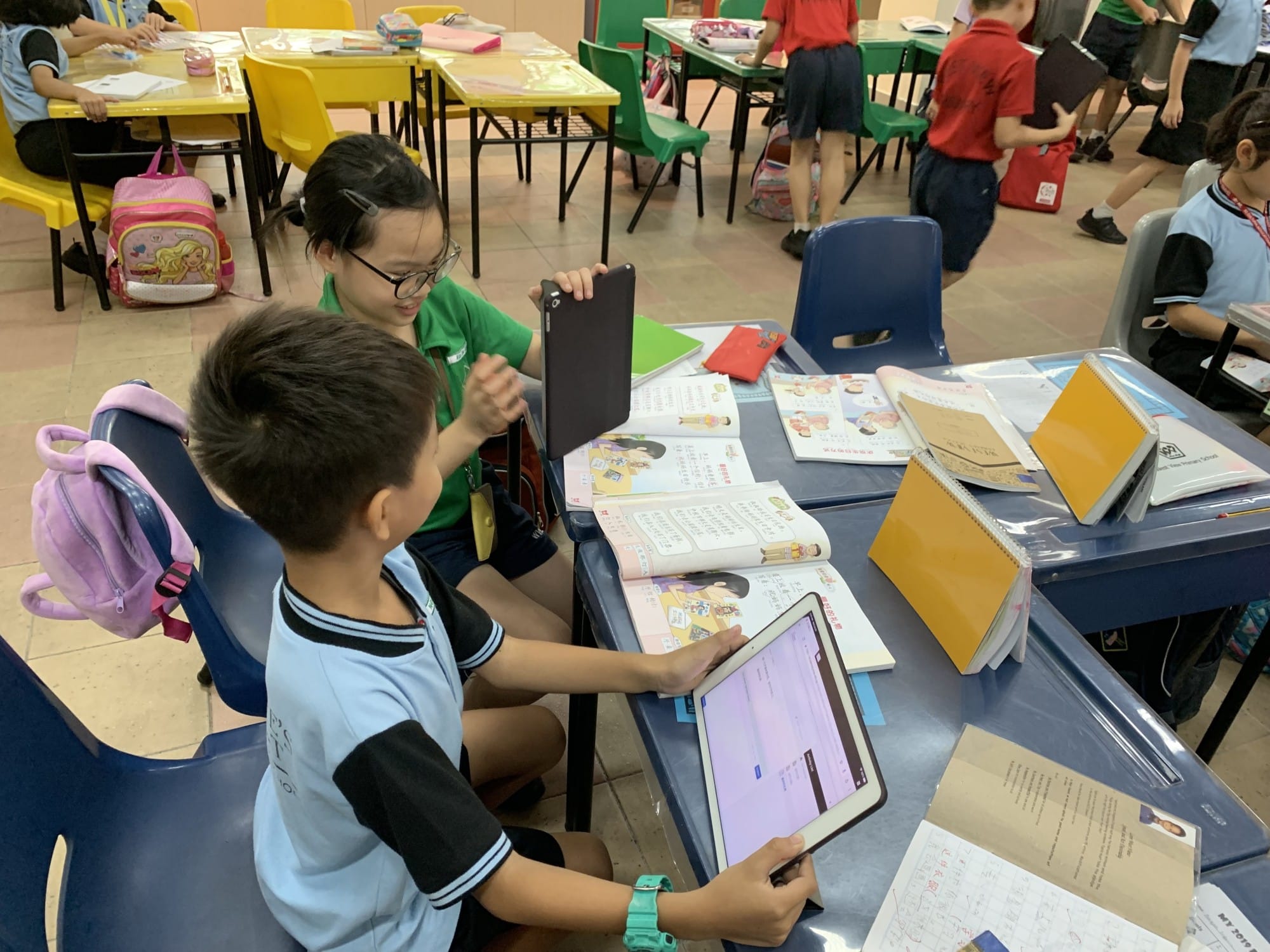 Two young students in school uniforms use tablets at a desk covered with open textbooks in a busy classroom.