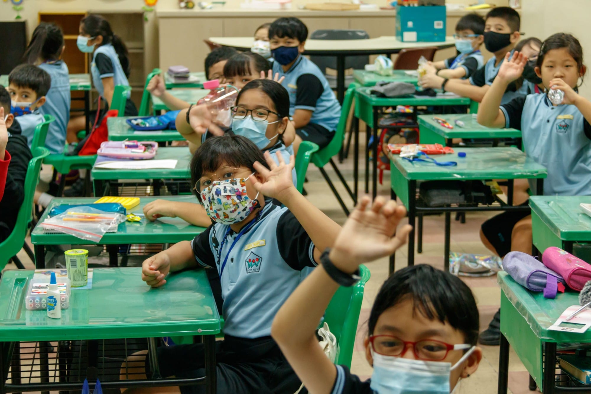 Young students in blue uniforms and face masks sit at green desks in a classroom, some raising their hands.