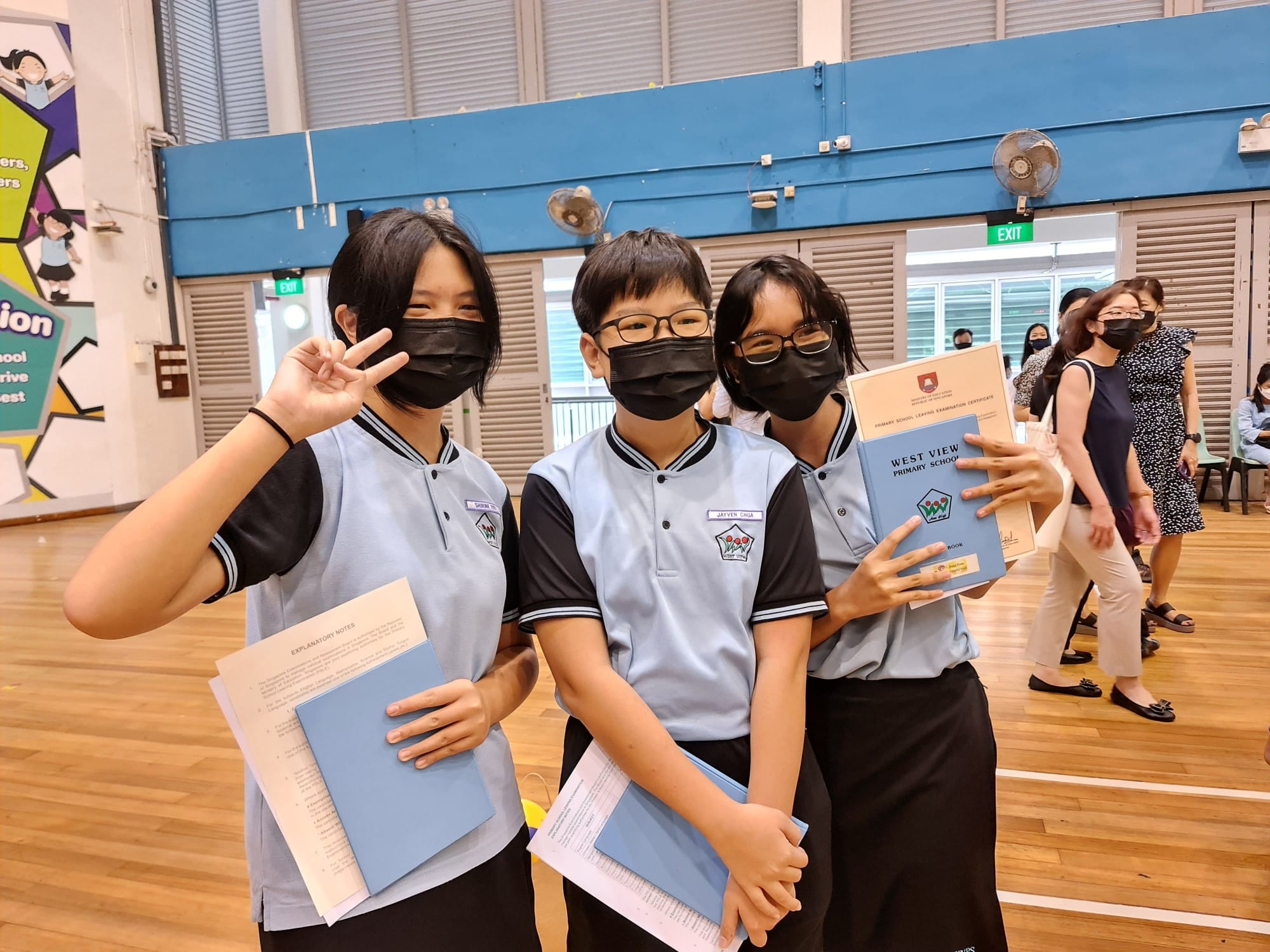 Three masked students in West View Primary School uniforms pose in a hall, holding certificates and documents.