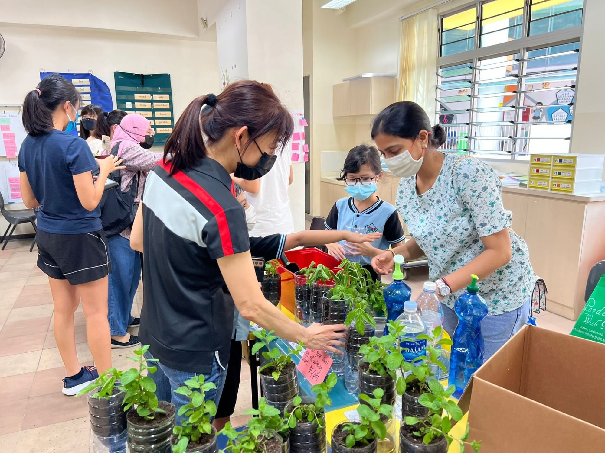 People wearing masks gather around a table with small potted plants and water bottles in a classroom setting.