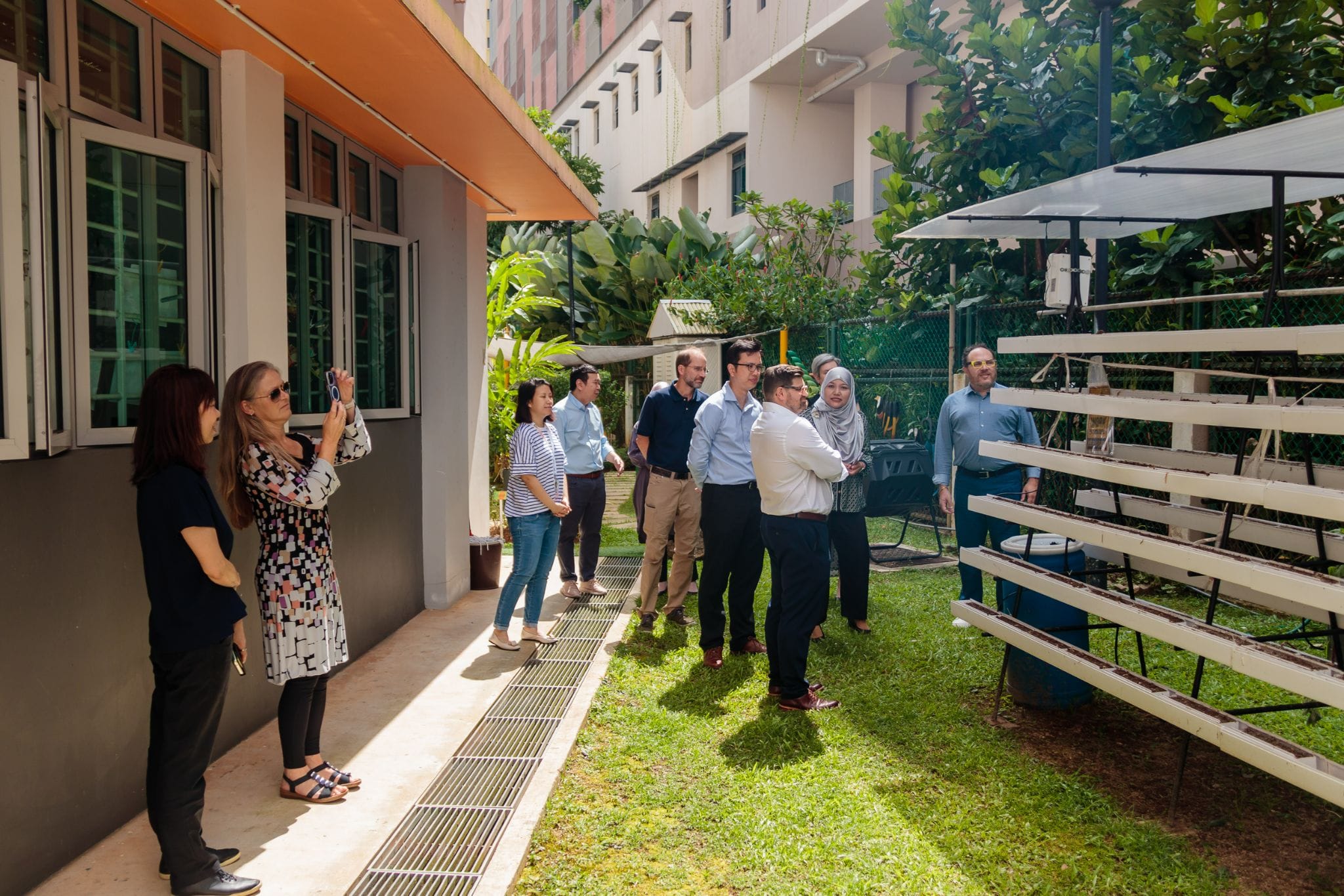 A group of people stand outdoors near a building, looking at tiered hydroponic growing racks on a green lawn.
