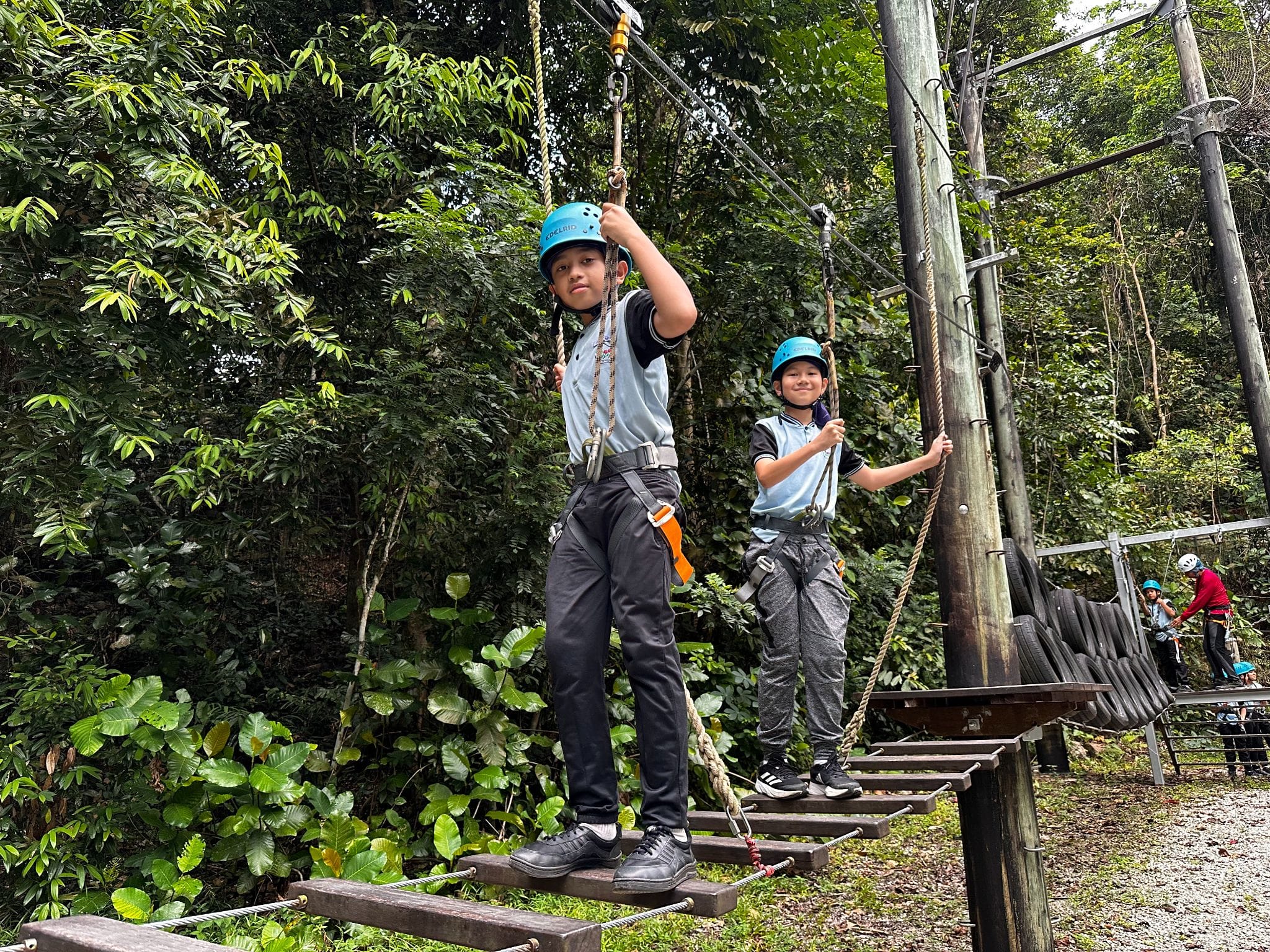 Two children wearing blue helmets and harnesses navigate a rope obstacle course surrounded by green trees.