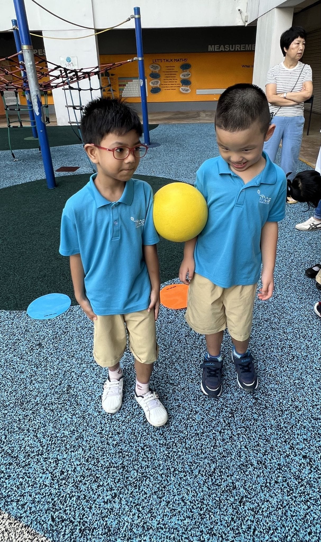 Two young boys in matching blue school uniforms hold a yellow ball on a playground with rubber flooring.