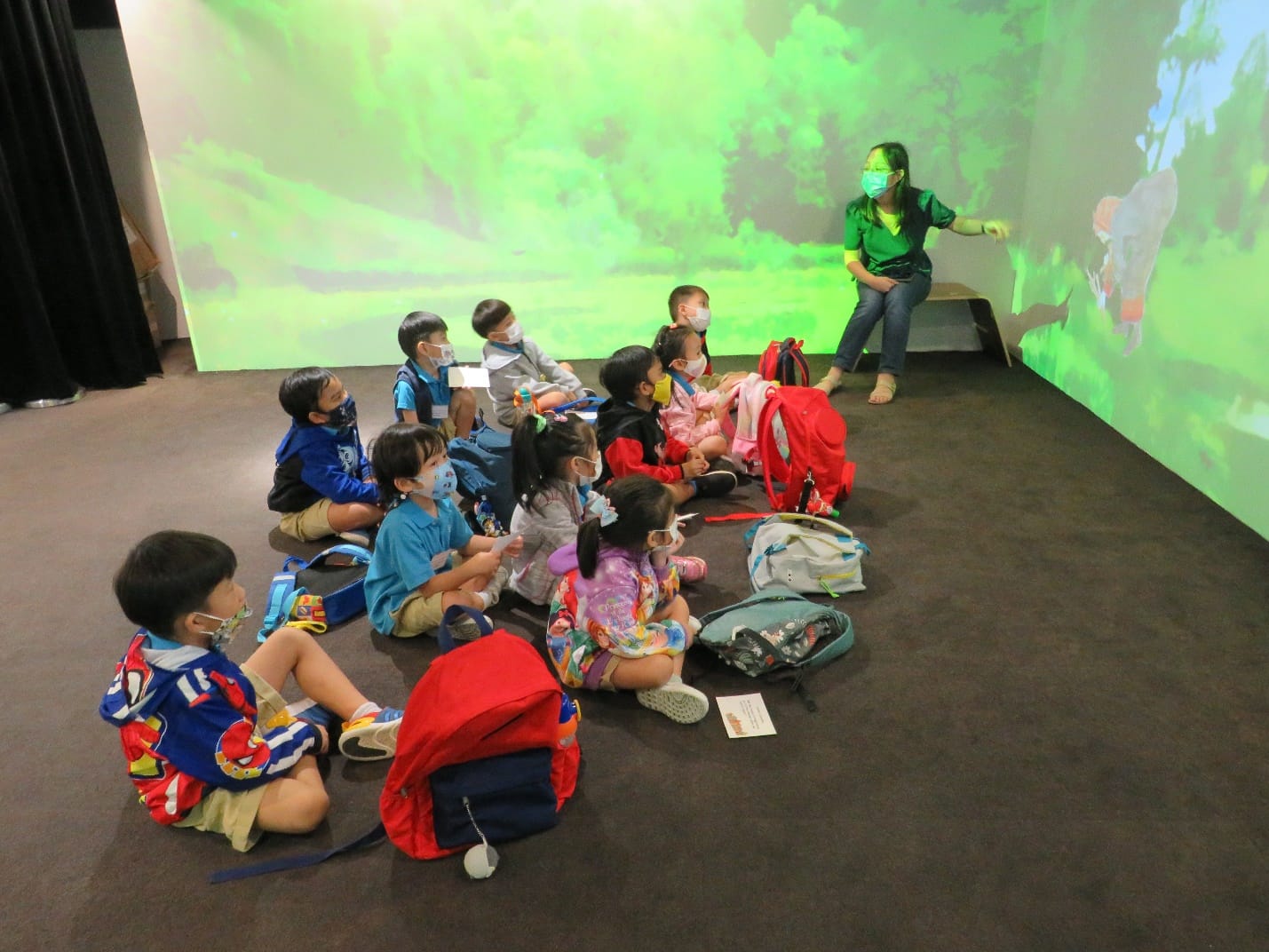 Young masked children sit on the floor facing a masked teacher gesturing at a green forest projection on the wall.
