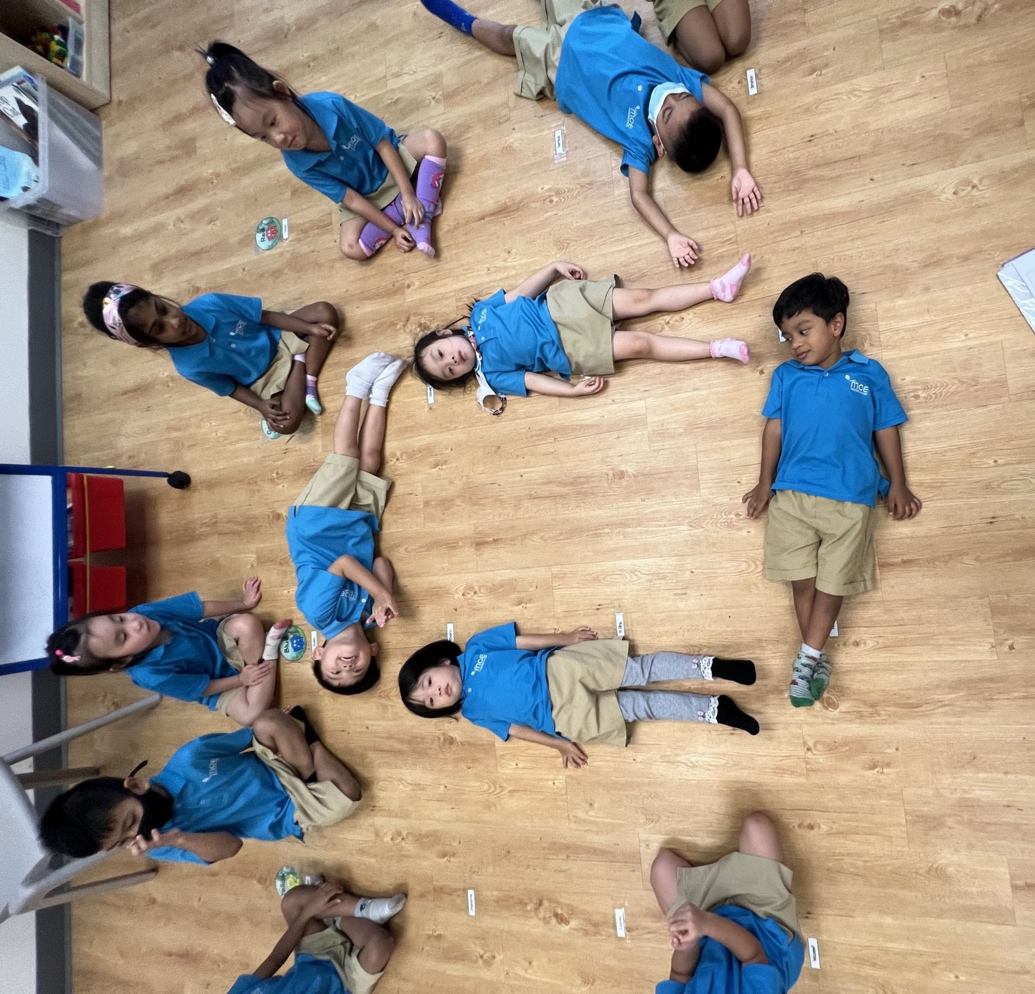 Several young children in matching blue and tan uniforms sit and lie on a wooden classroom floor.