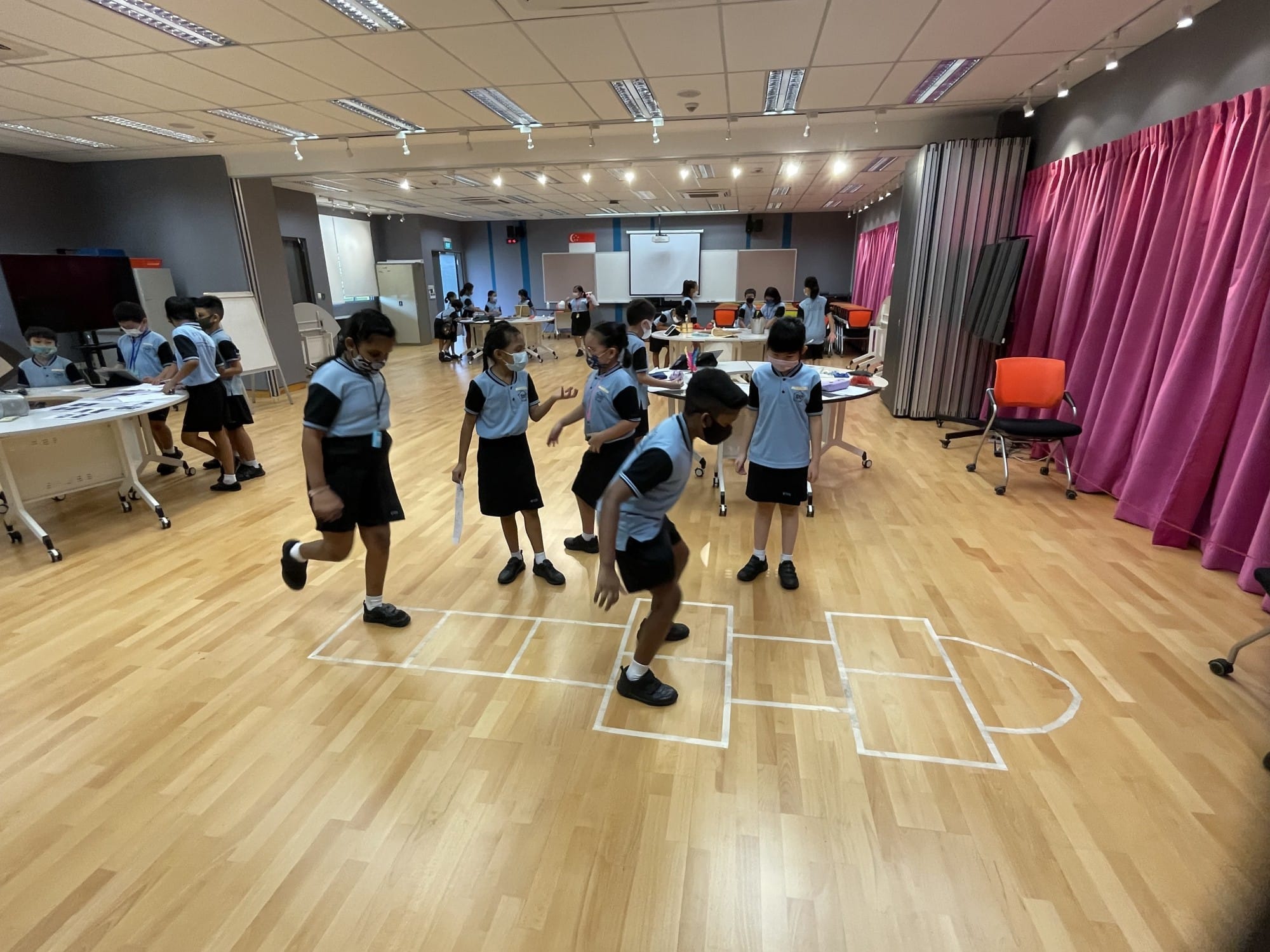 Children in school uniforms and masks play on a wooden floor with taped shapes in a bright indoor hall.