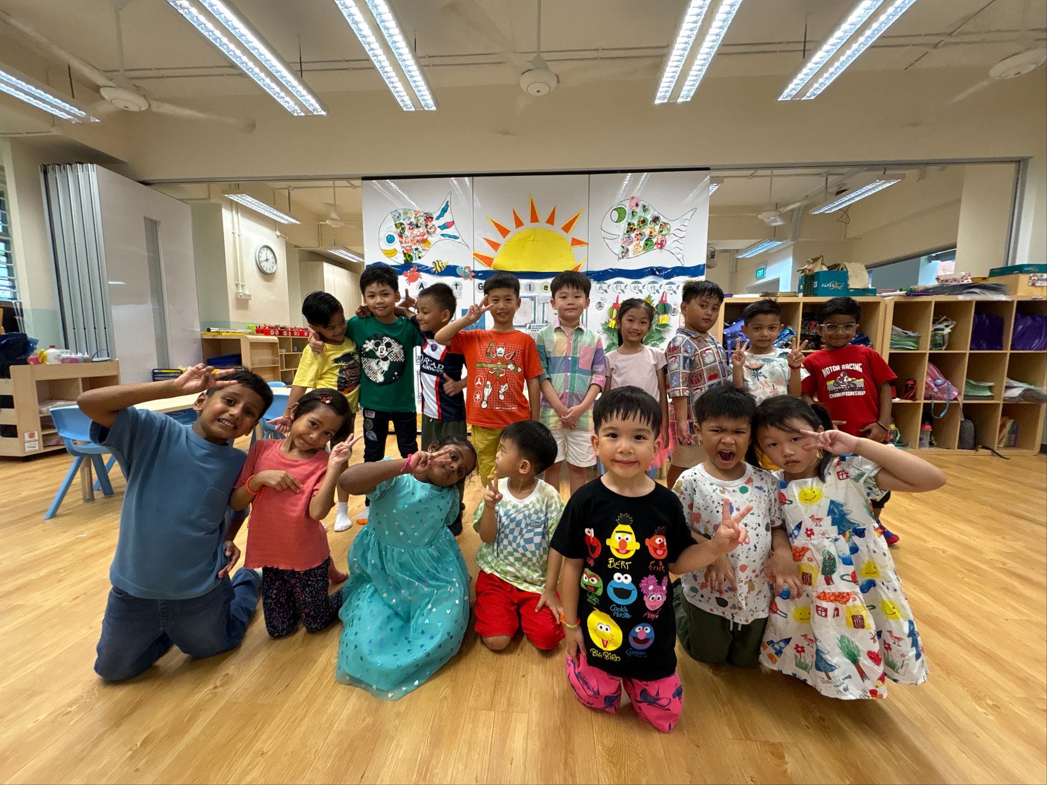 A group of young children posing and smiling in a bright classroom with a colorful sun and fish mural behind them.