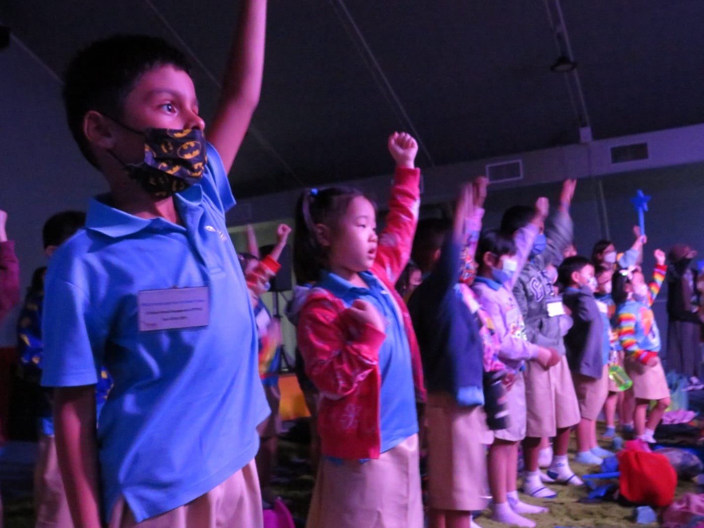 Children in blue uniforms raise their hands enthusiastically in a dimly lit room, some wearing face masks.