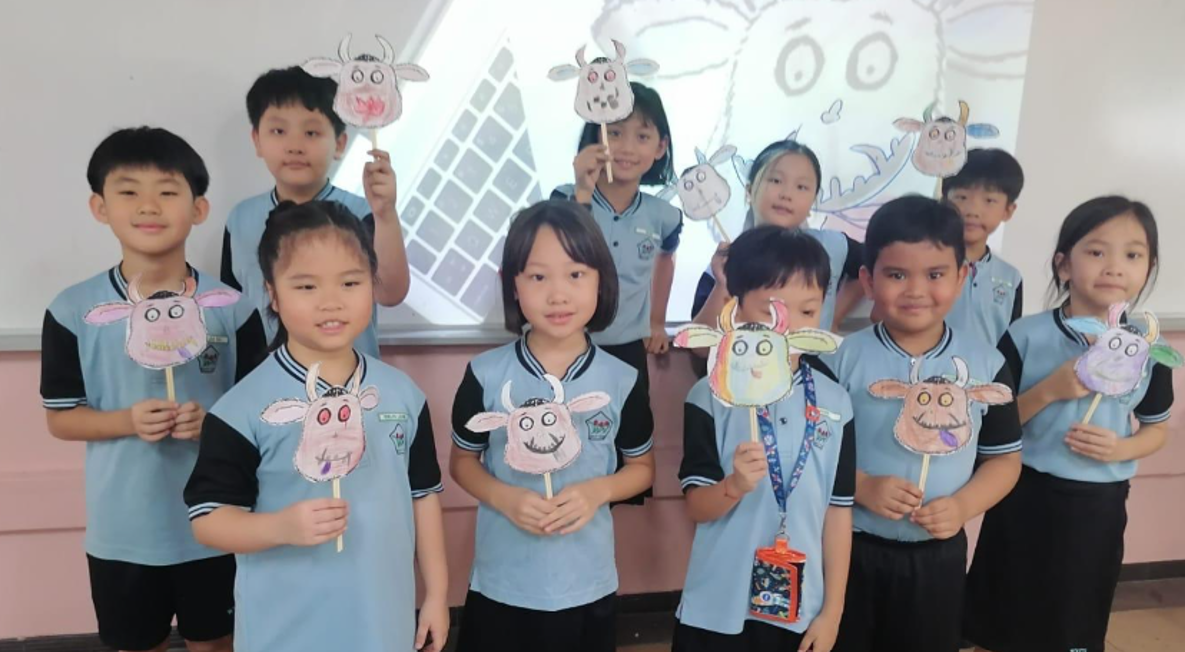 Ten children in blue uniforms hold hand-made paper cow face puppets on sticks in a classroom.