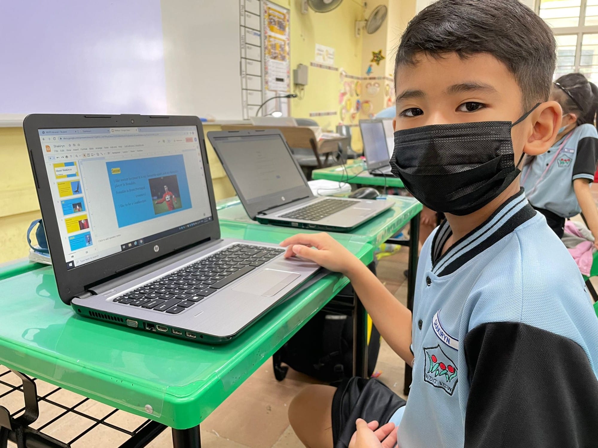 Young student wearing a black face mask sits at a green desk using an HP laptop in a classroom.