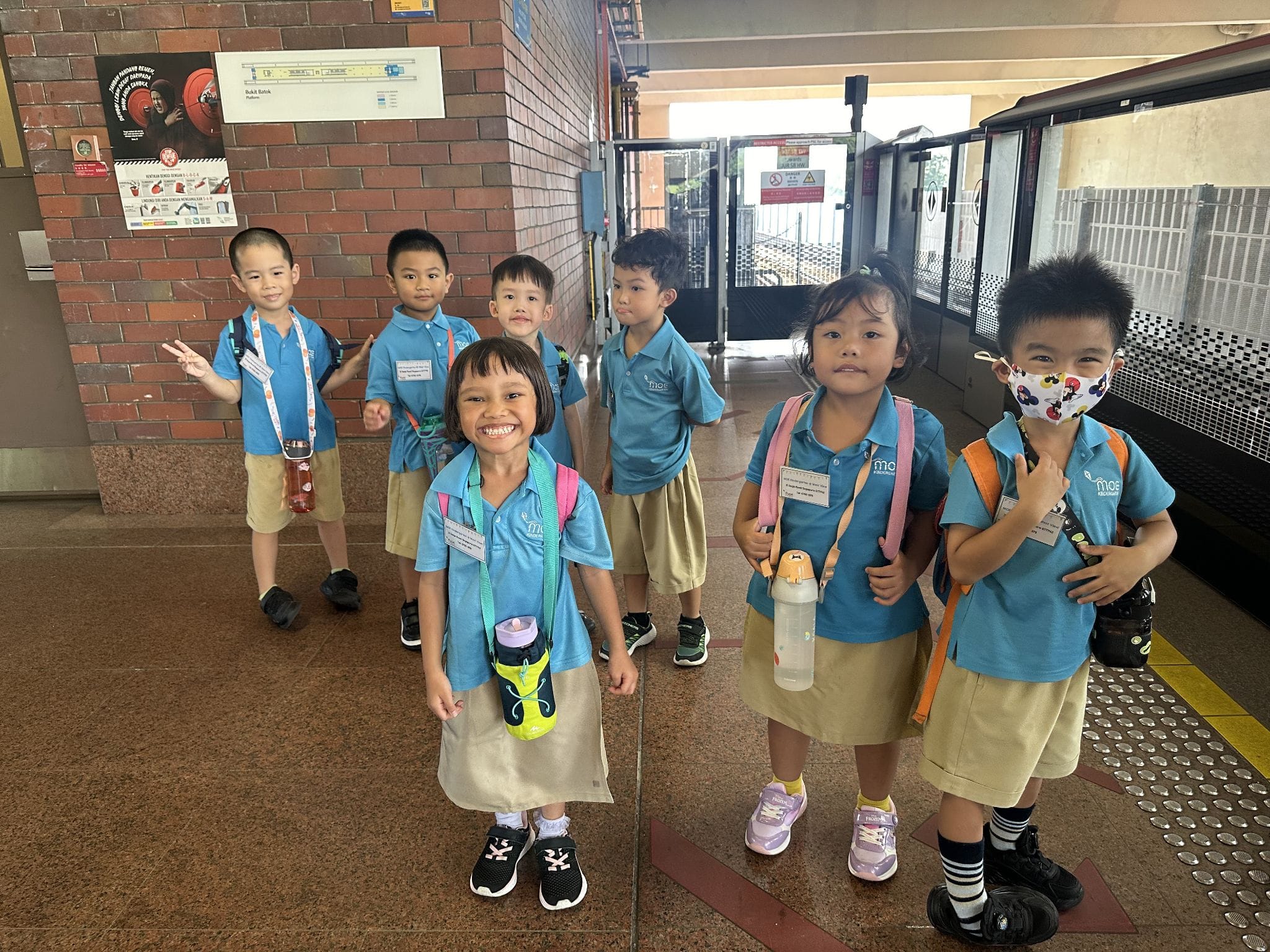 Seven young children in matching blue uniforms and backpacks stand smiling at a train station platform.