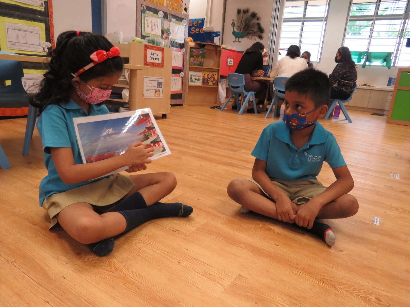 Two masked children in blue uniforms sit cross-legged on a classroom floor, with a girl holding a photo toward a boy.