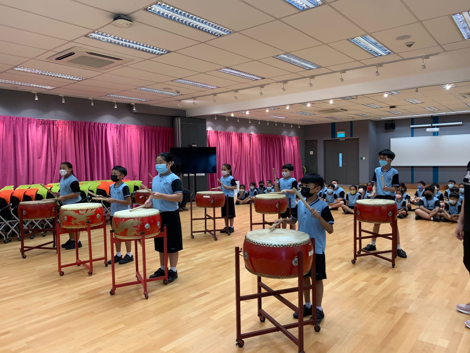 Children in school uniforms and masks play red Chinese drums on a wooden floor with pink curtains behind them.