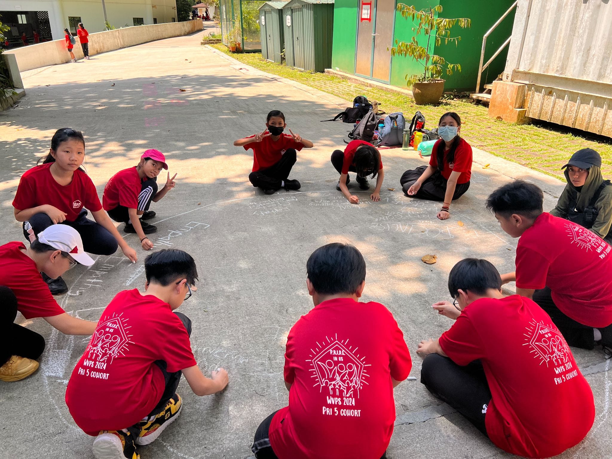 Children in red WWPS 2024 Pri 5 Cohort shirts sit in a circle on pavement, writing with chalk outdoors.