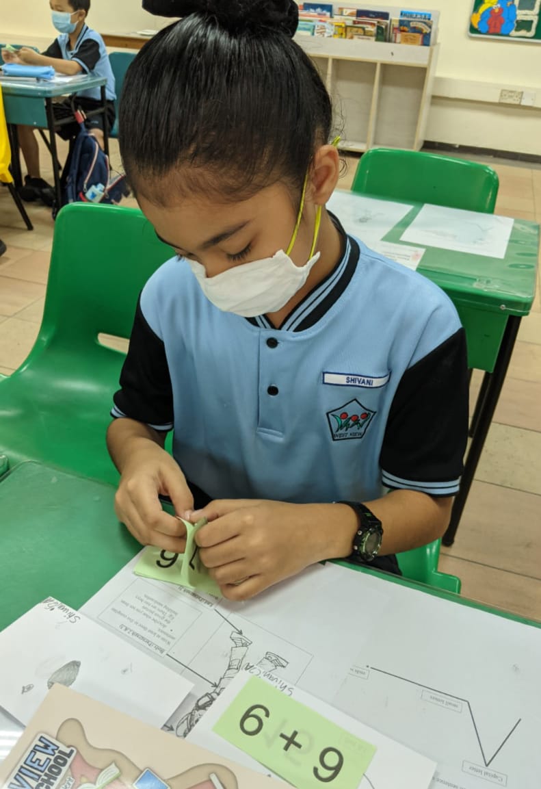 A young student named Shivani in a blue uniform and face mask works on a math addition worksheet showing 6 plus 9.