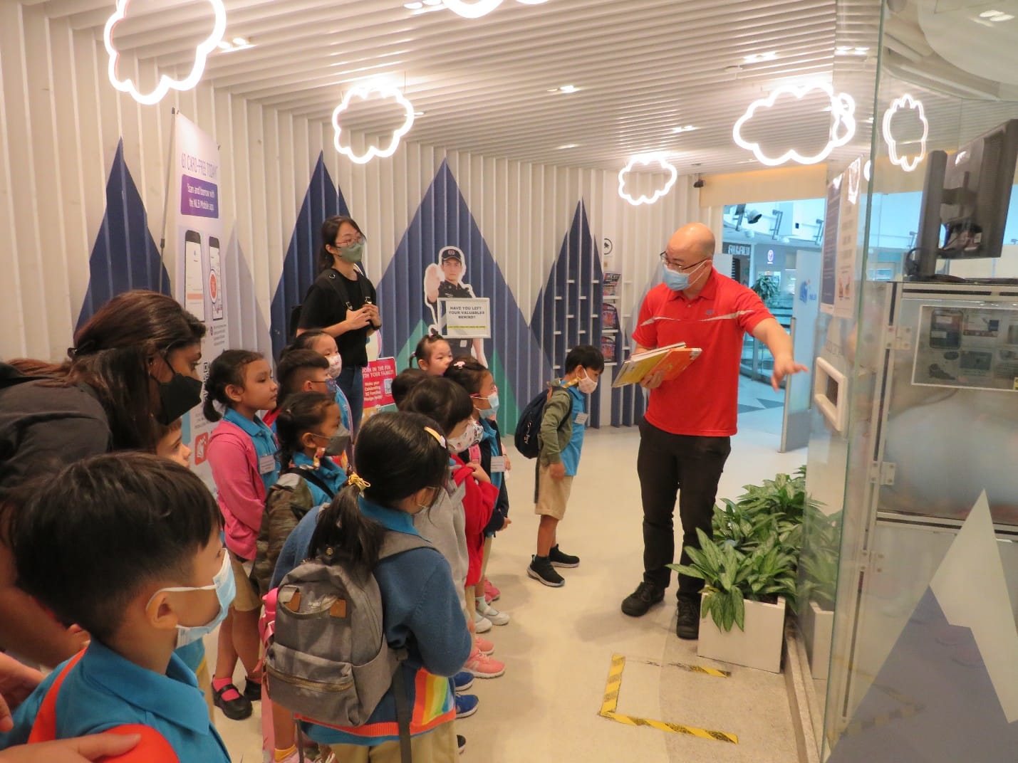 A man in a red shirt wearing a mask gestures to a display while young children on a field trip watch attentively indoors.
