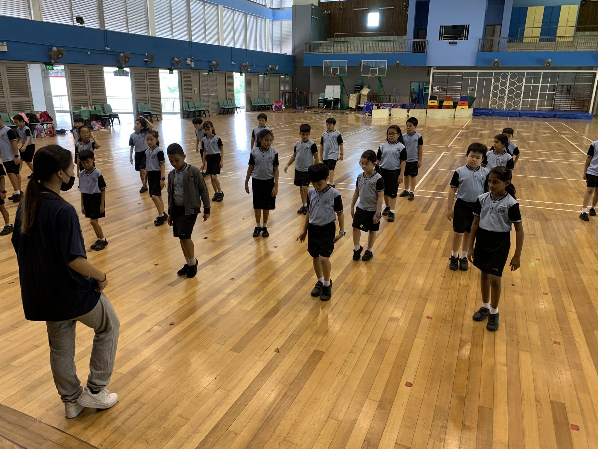 A teacher leads a large group of young students in uniforms during a PE class inside a school gymnasium.