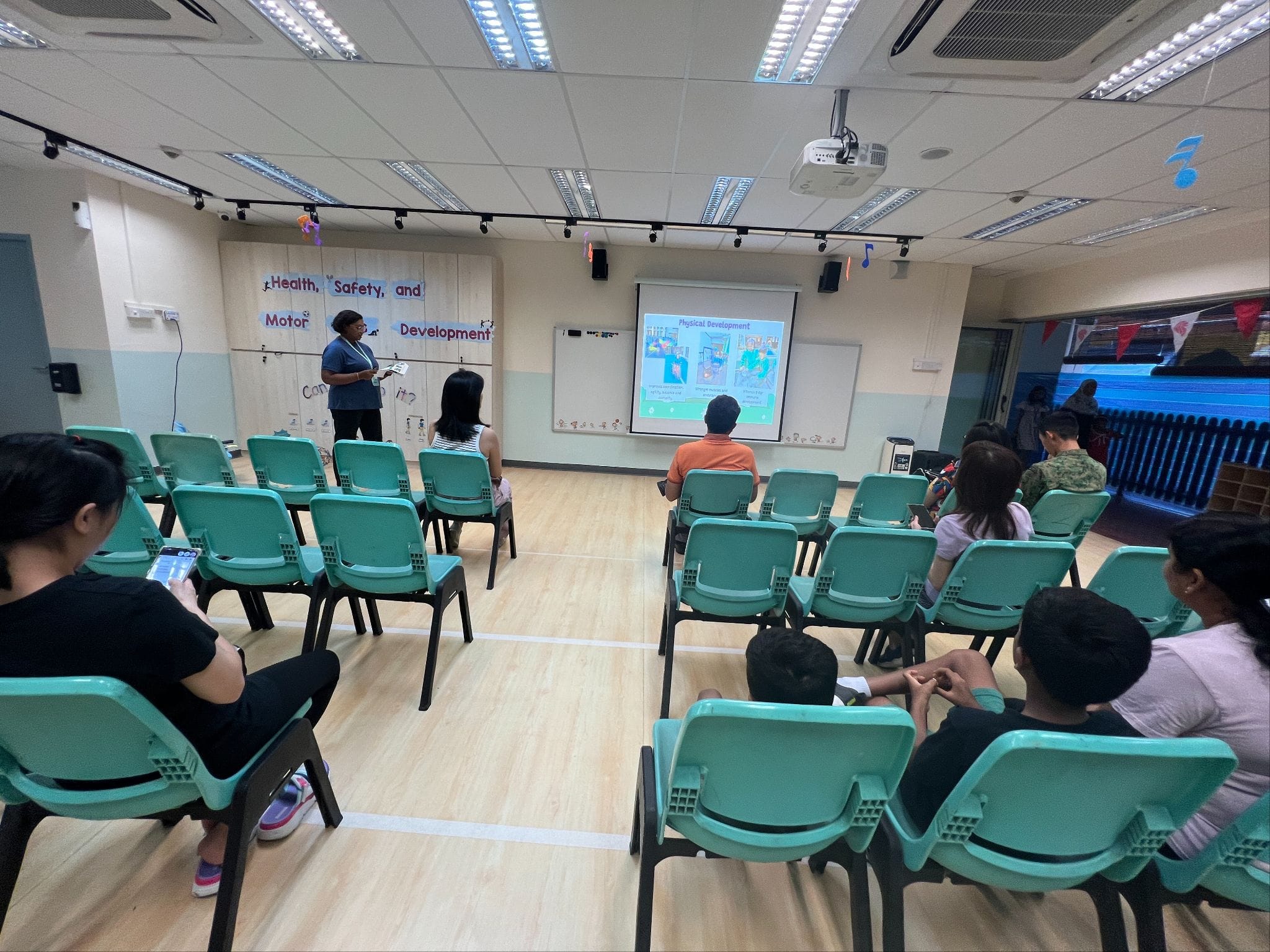 A presenter stands at the front of a classroom with teal chairs and a projected slide on Health, Safety, and Motor Development.