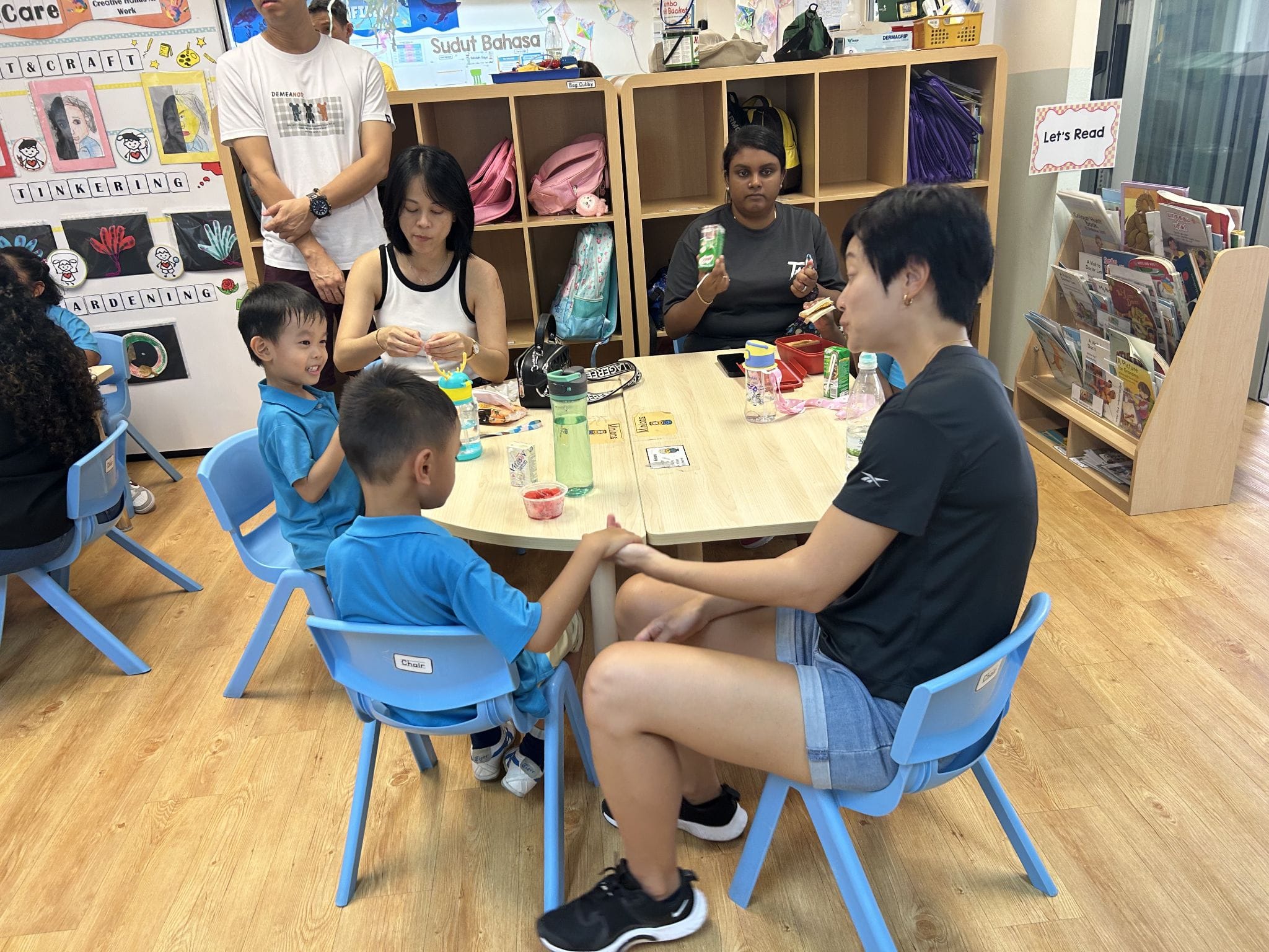 Adults and young children in blue uniforms sit around a small table in a colorful classroom with bookshelves.