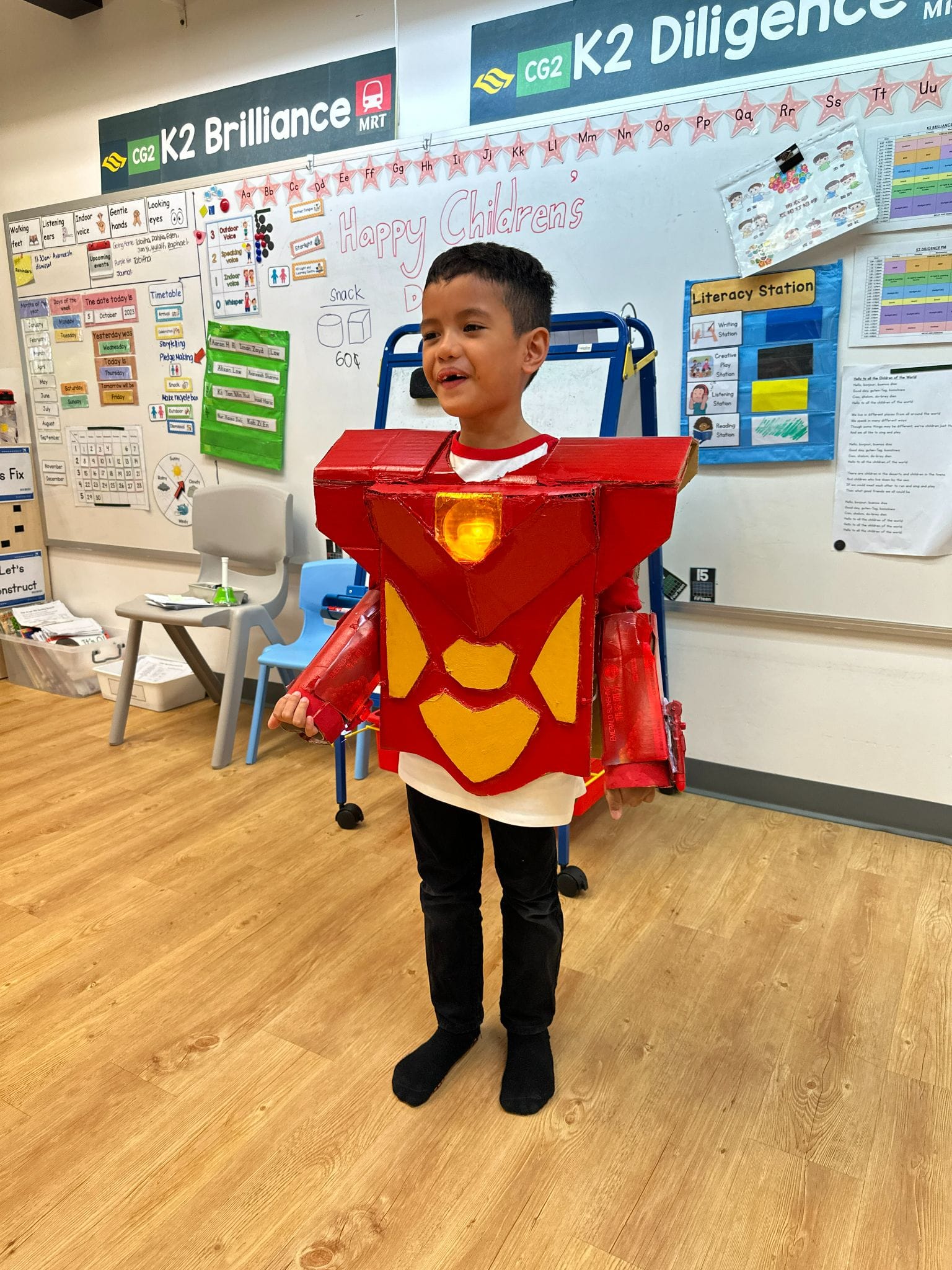 A young boy wearing a red and yellow cardboard Iron Man costume stands smiling in a decorated classroom.