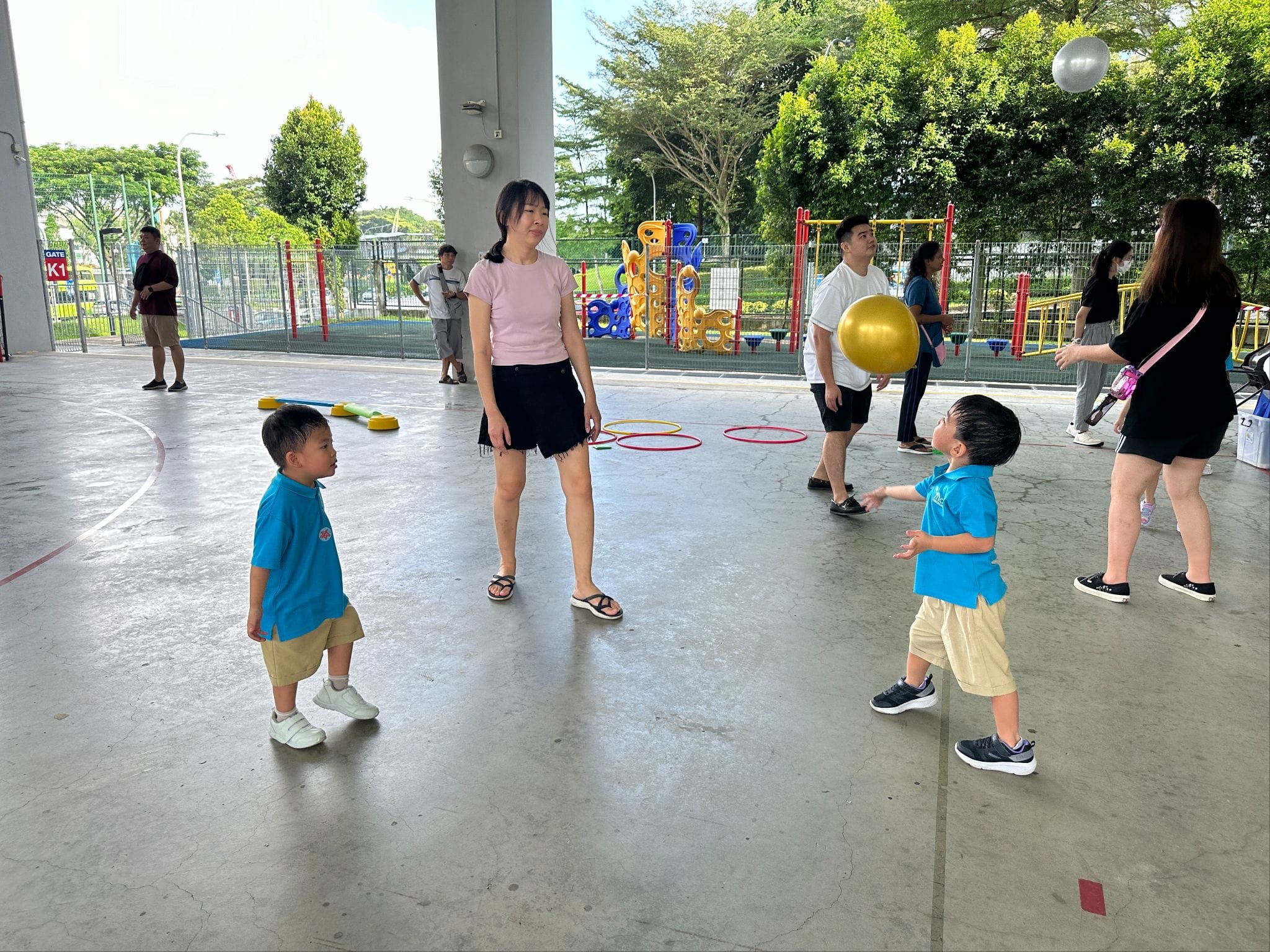 Two young children in blue shirts play with balloons near adults on an outdoor covered concrete court with colorful hoops.