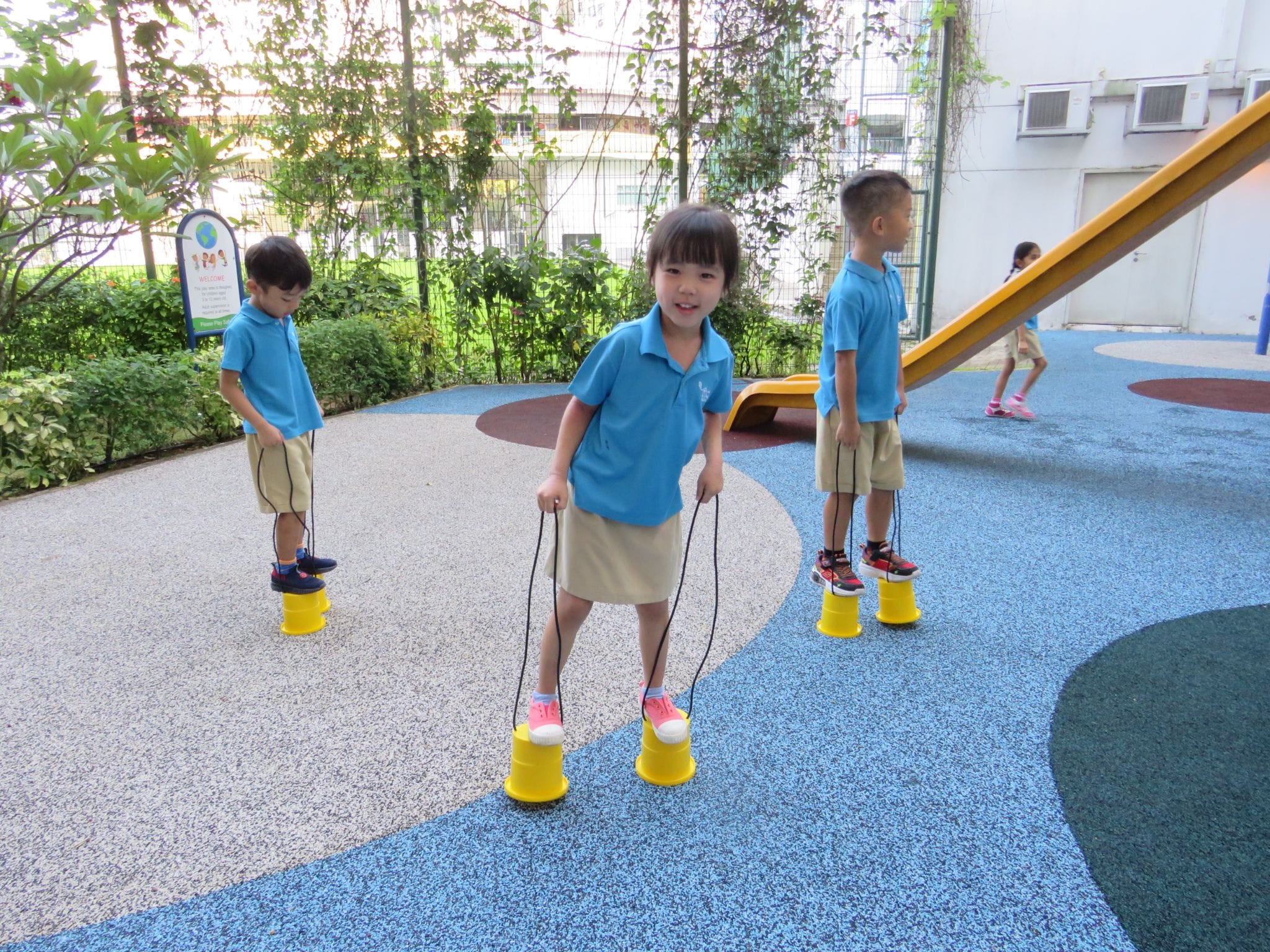 Three children in blue uniforms balance on yellow cup stilts with strings on a colorful playground.