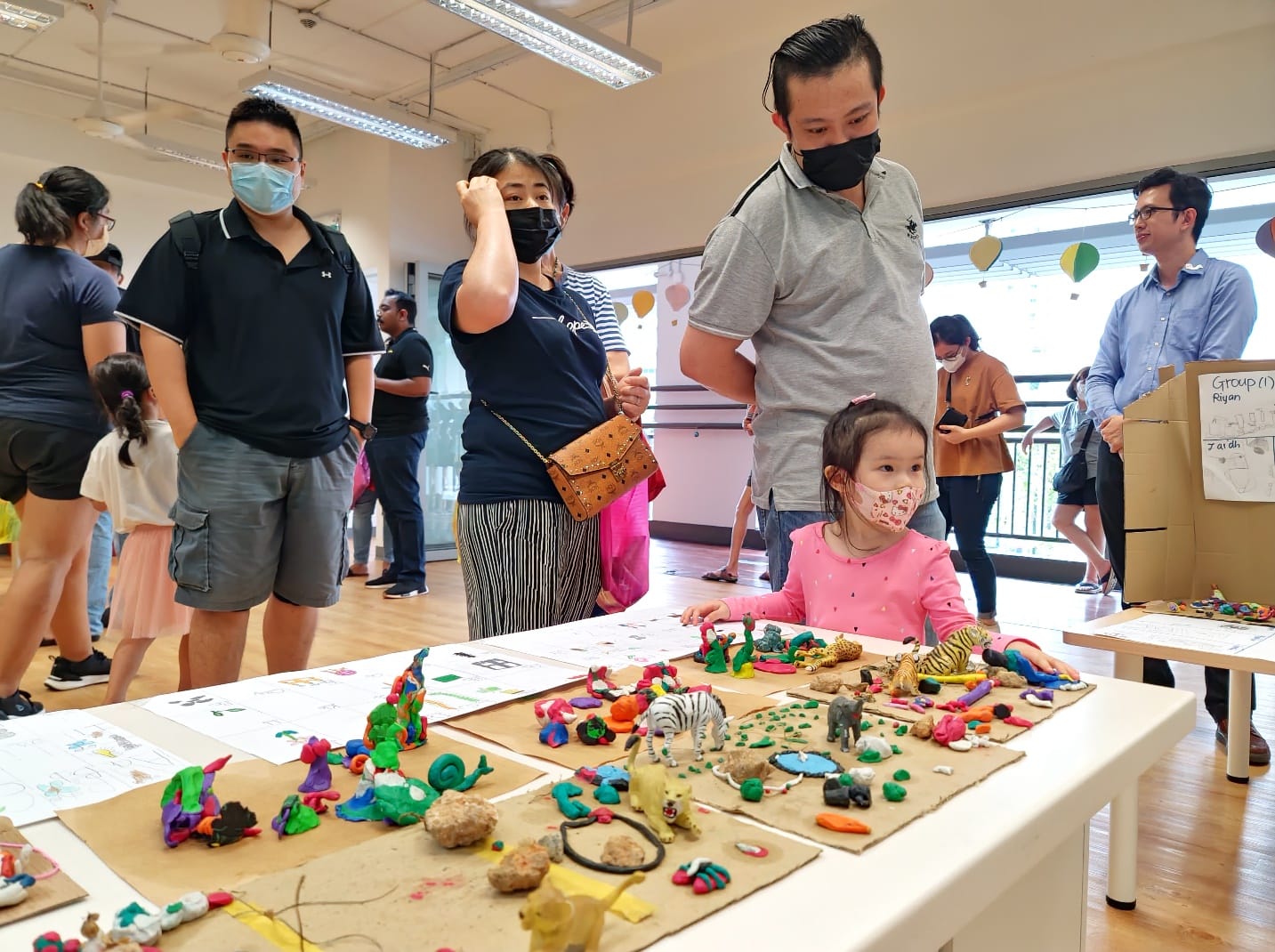 A young girl and masked adults view a table displaying colorful clay sculptures and toy animals at an indoor event.
