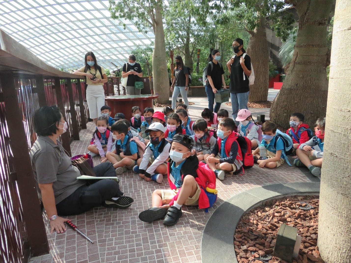 A masked adult guide addresses a large group of masked young children sitting on a brick path inside a greenhouse.