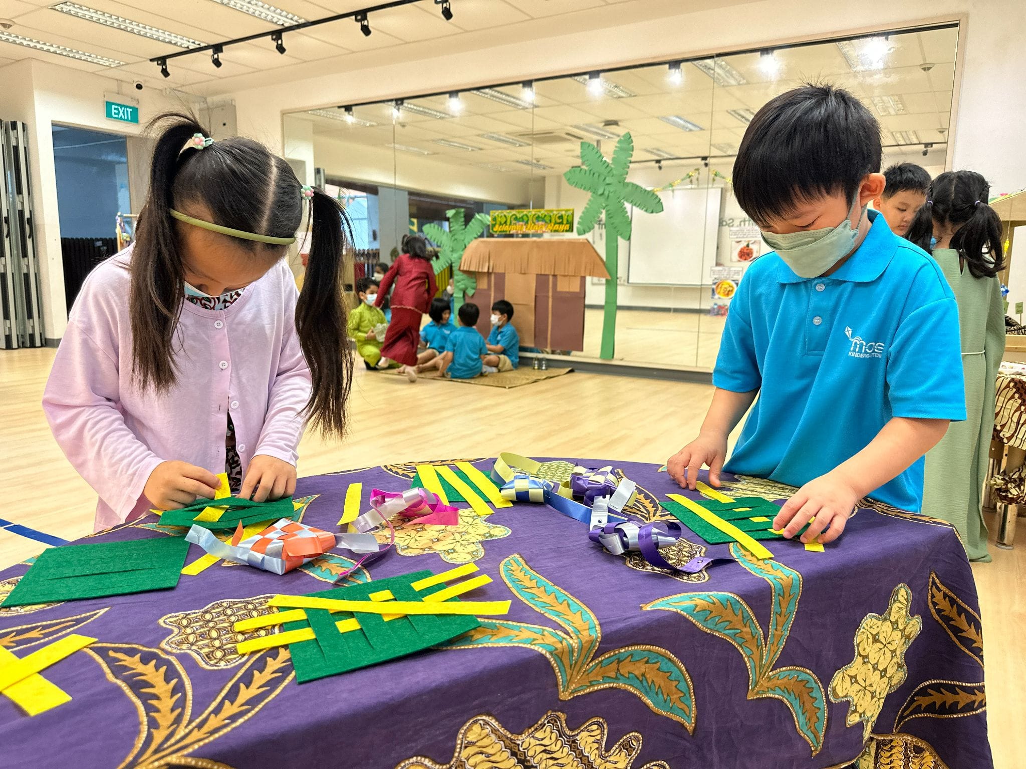 Two young children wearing masks weave colorful paper strips at a table covered with a decorative purple cloth.