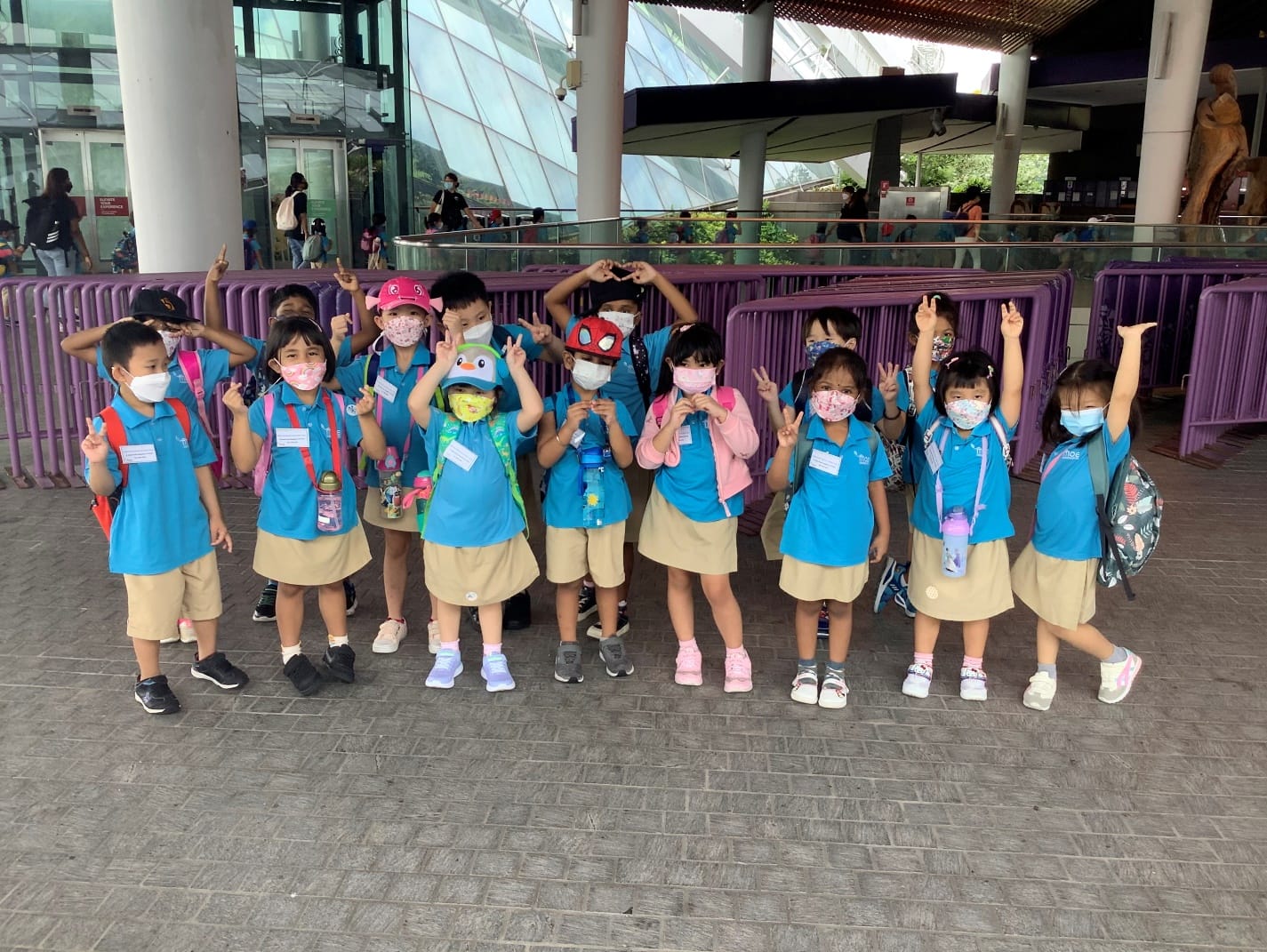 Group of young children in blue uniforms and face masks posing with peace signs outside a modern building.